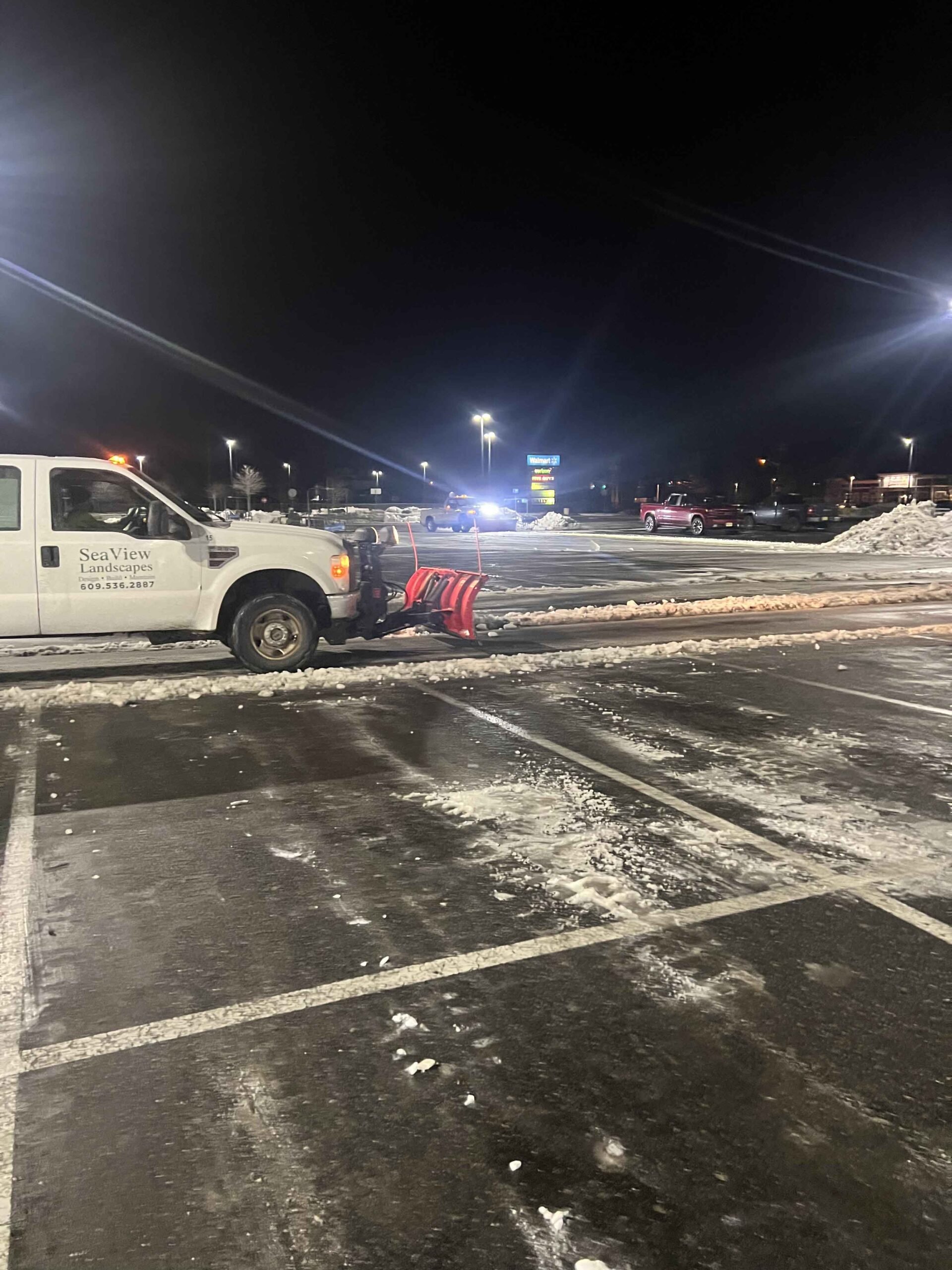 A SeaView Landscapes truck with a red front-mounted plow clears a commercial parking lot at night after a snowstorm. Snow is pushed to the lot edges and lot markings are visible on the wet asphalt.