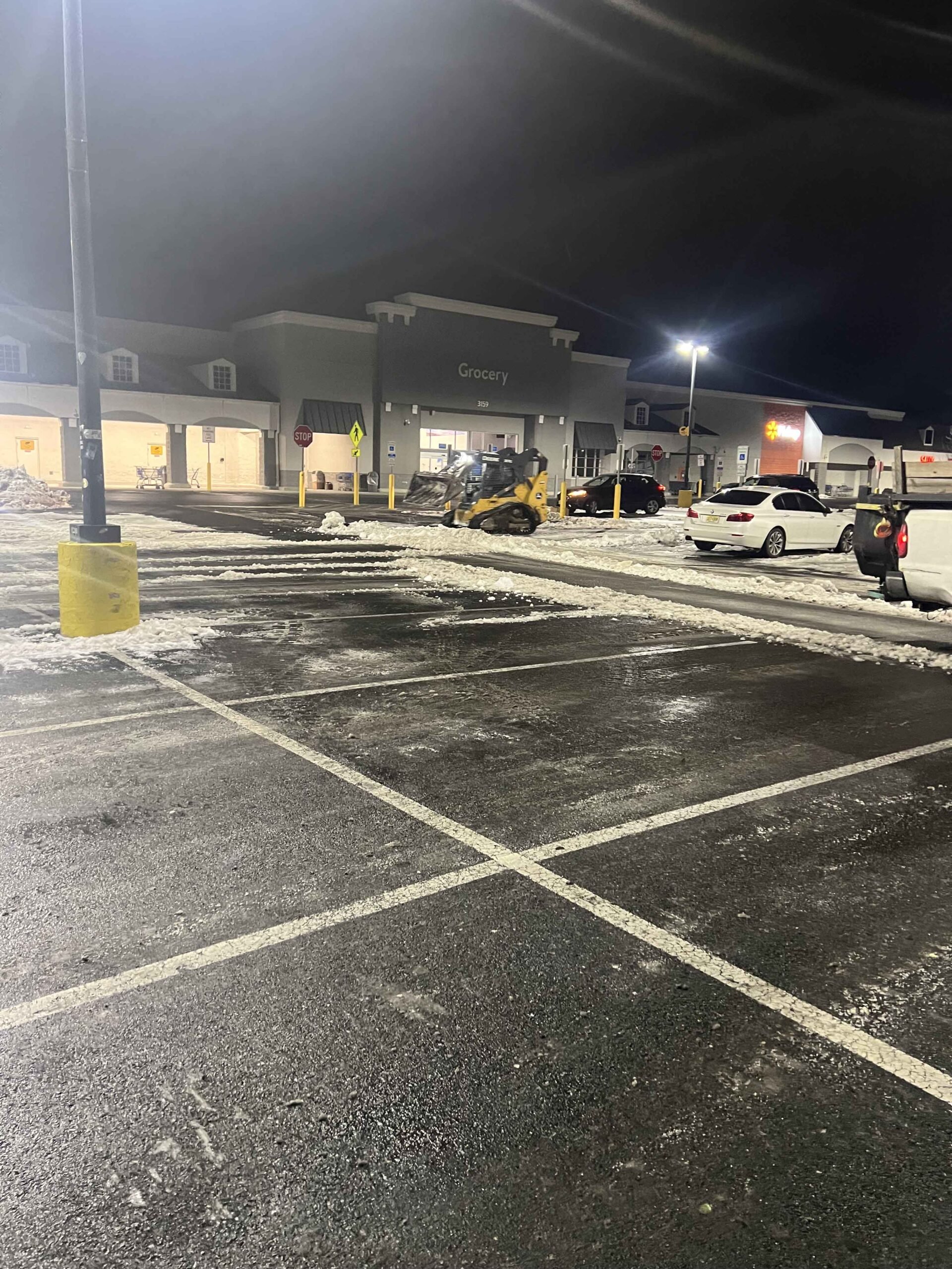 A skid-steer loader clears snow from a commercial asphalt parking lot at night in front of a grocery store. The lot surface is wet with residual slush and snow piles are visible at the lot perimeter. Overhead parking lot lights illuminate the work area.