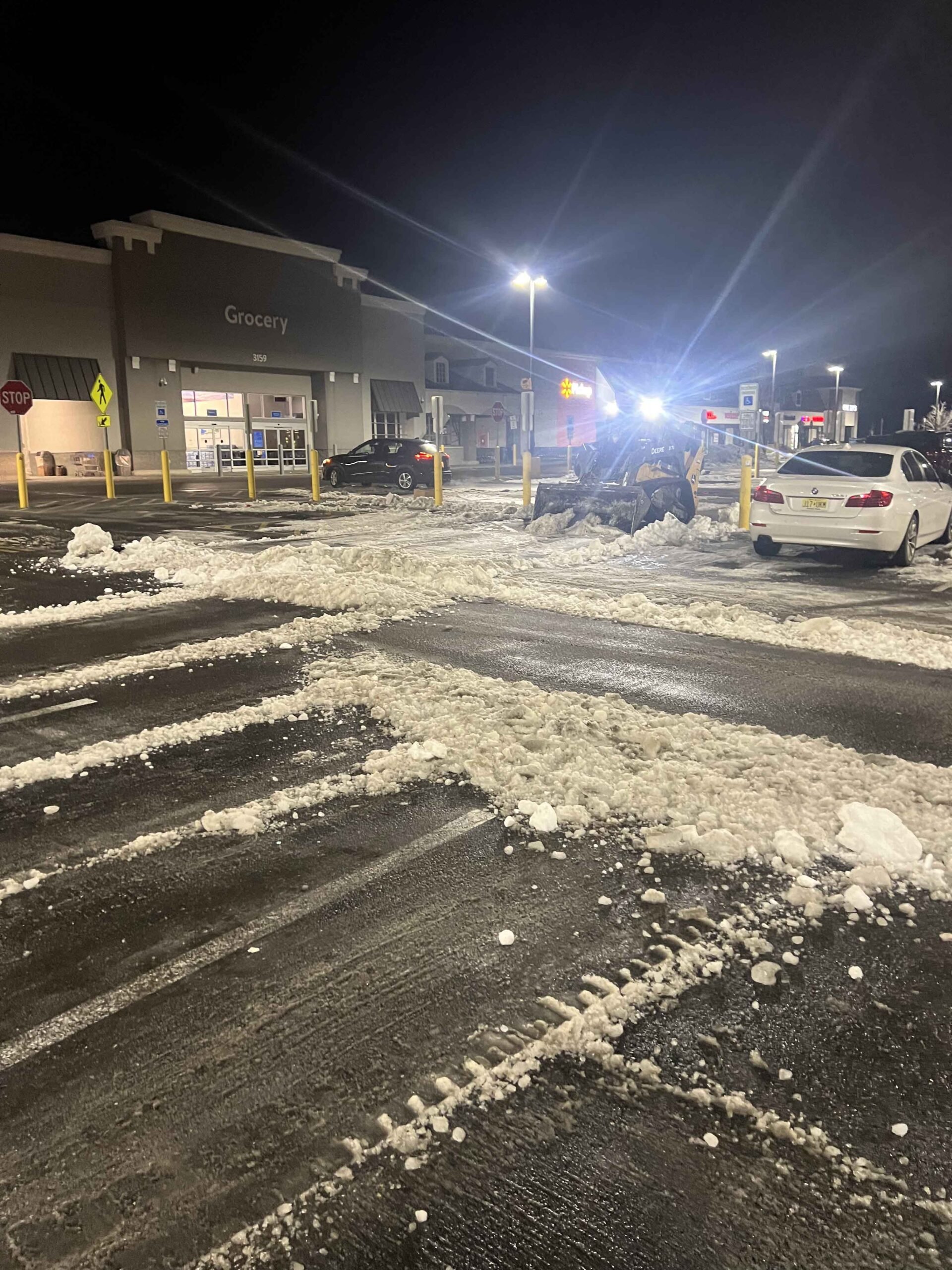 A commercial grocery store parking lot is being plowed at night, with snow piled along the driving lanes and a piece of equipment visible mid-lot. The asphalt surface is wet and cleared of accumulated snow, with overhead parking lot lights illuminating the