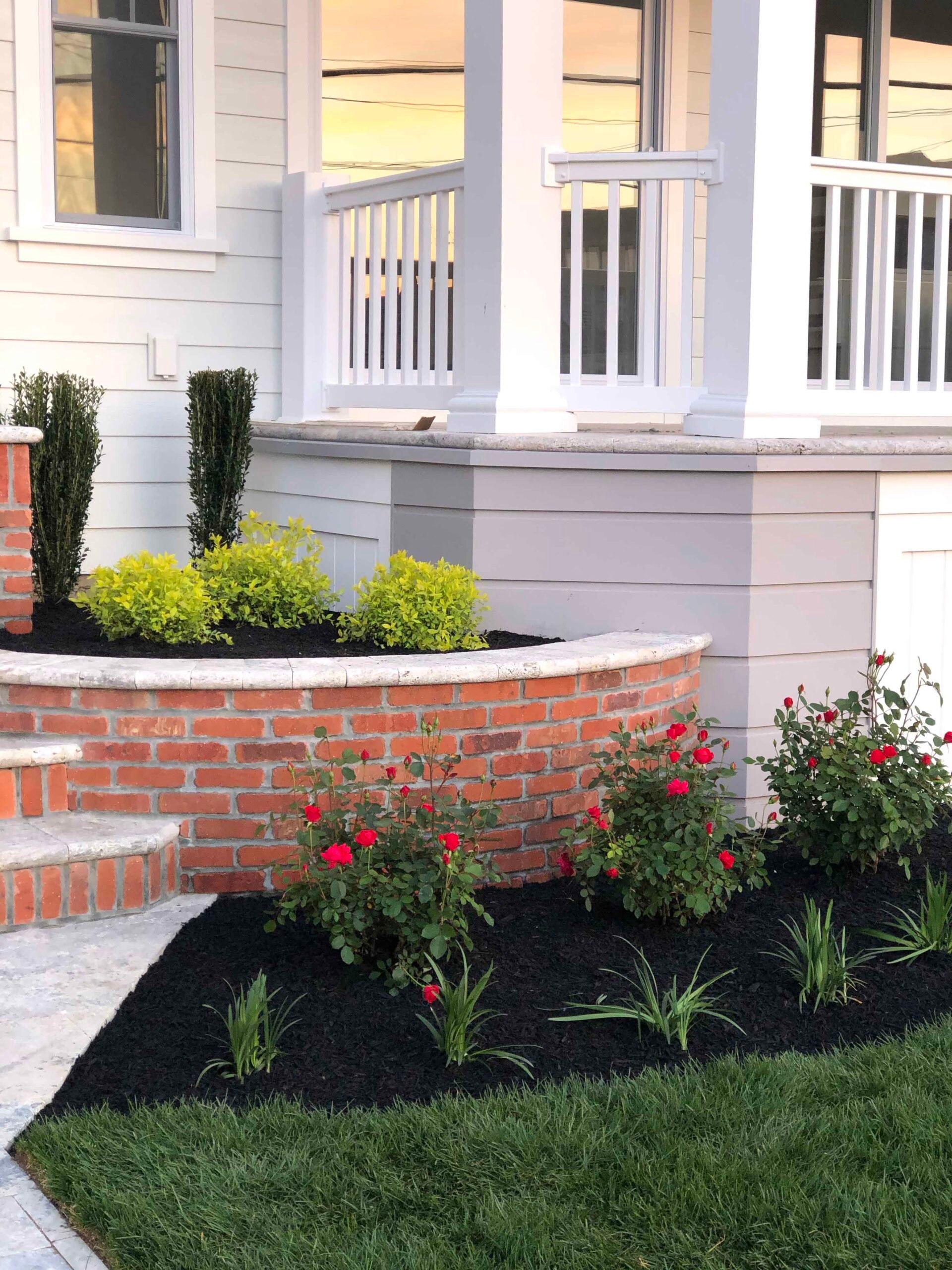 A curved brick retaining wall borders a raised planting bed filled with black mulch, red knockout roses, ornamental grasses, and upright evergreen shrubs adjacent to a porch foundation.