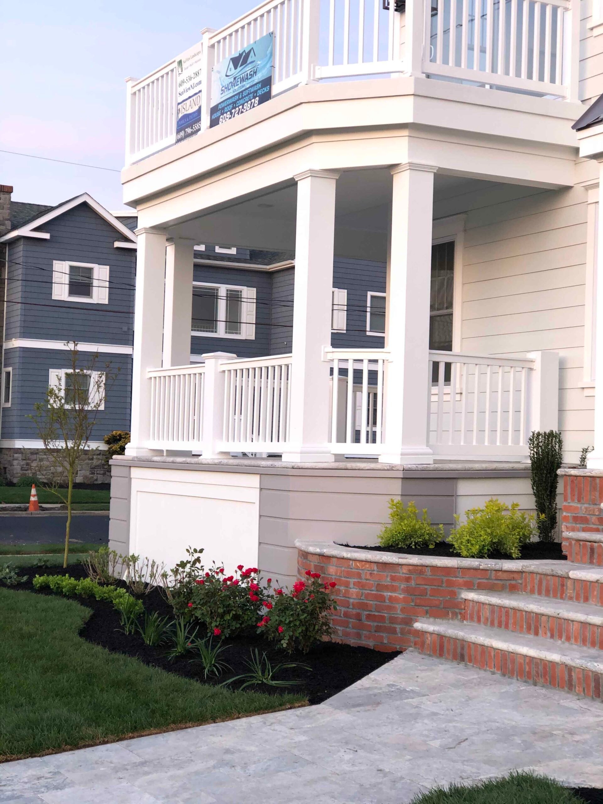 A front porch entry features a curved brick retaining wall with a new planting bed filled with dark black mulch, boxwood shrubs, red knockout roses, and ornamental grasses. A bluestone or concrete walkway leads to red brick entry stairs.