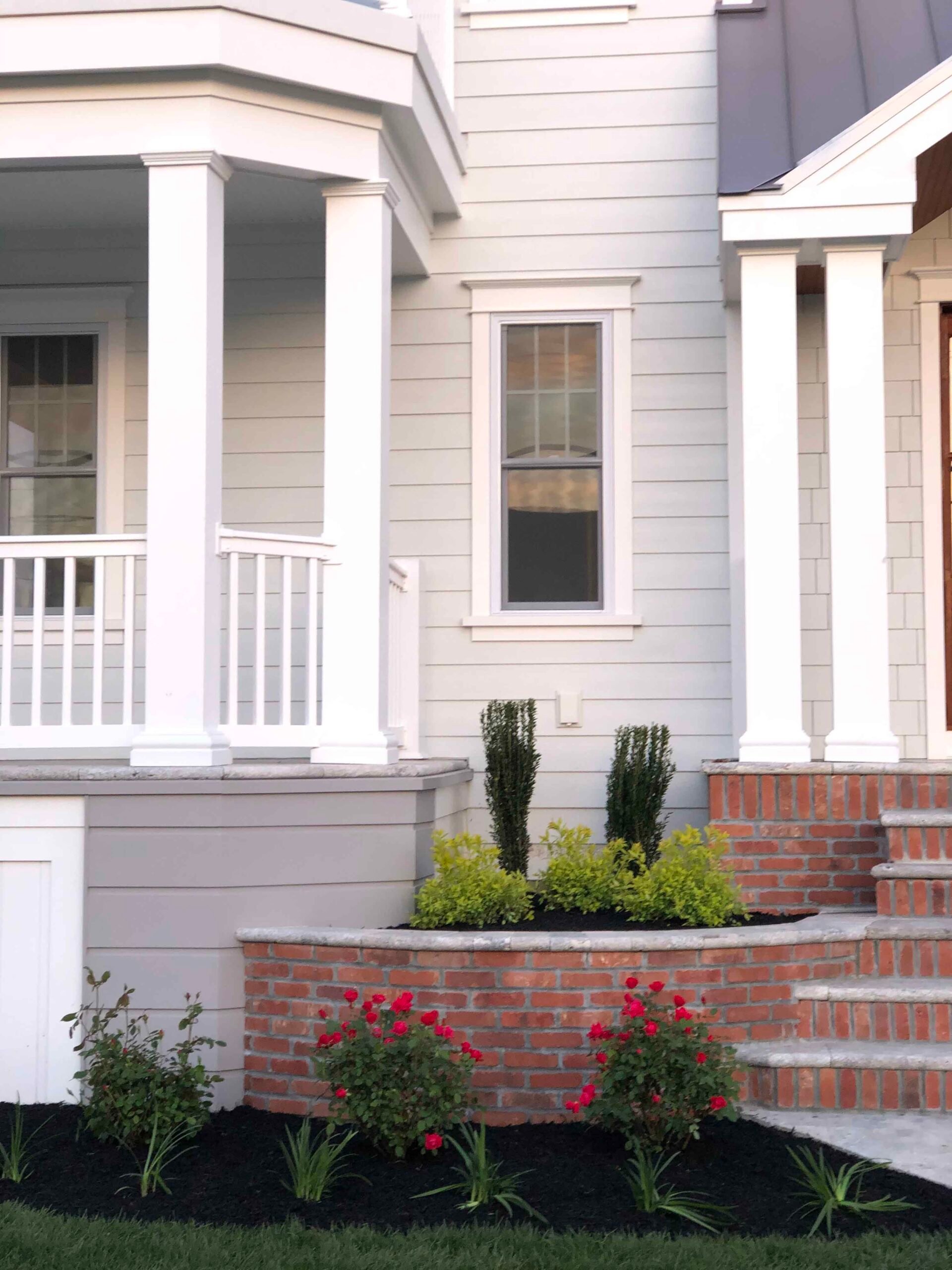 A curved raised brick planting bed fronts a gray coastal home, planted with compact upright evergreen shrubs, low yellow-green groundcover, and red flowering roses over black mulch. Brick steps with a bluestone cap lead to the front porch.