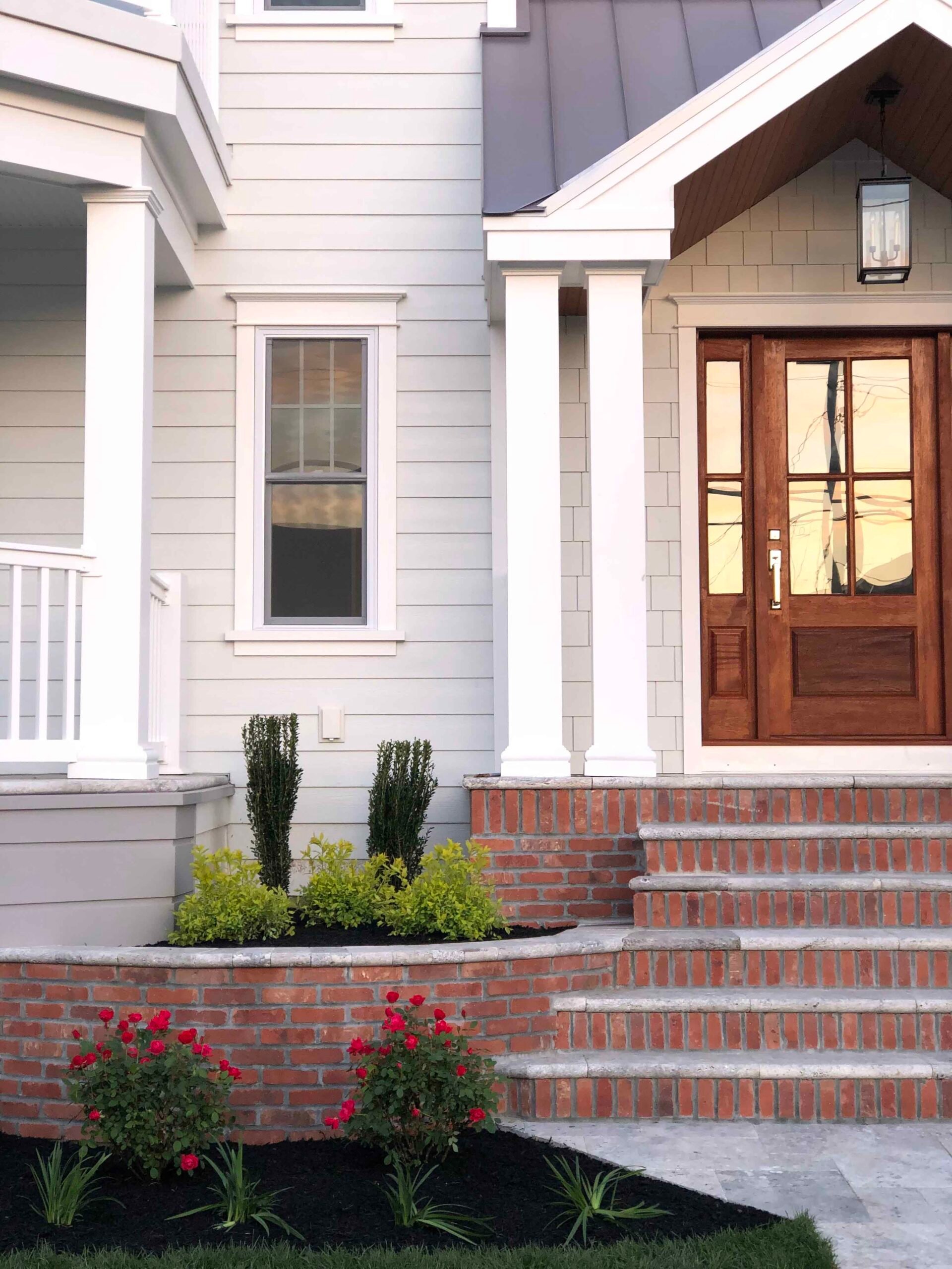 A curved brick retaining wall borders a freshly mulched planting bed at a home's front entrance, containing upright arborvitae, low boxwood-style shrubs, and red flowering roses. Dark black mulch covers the bed and bluestone pavers lead to the front steps.