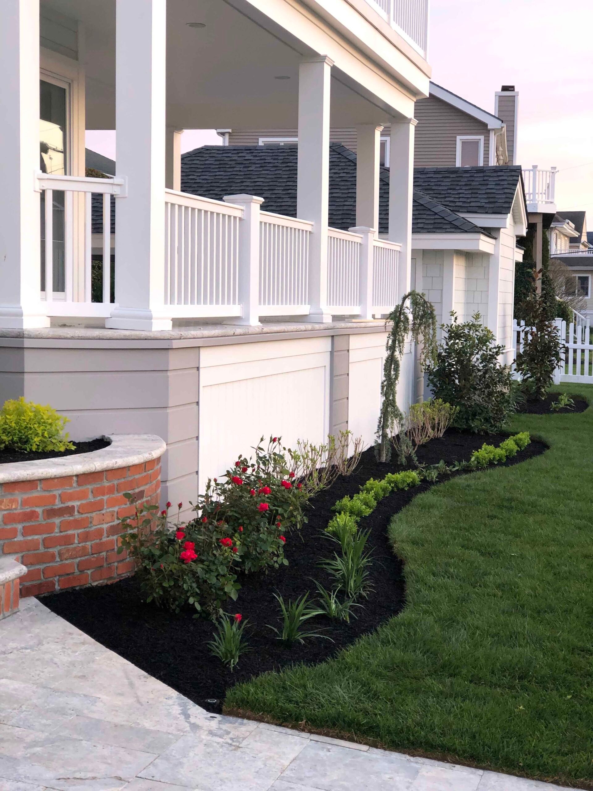 A curved front foundation planting bed runs along a residential porch, filled with red rose bushes, ornamental shrubs, and groundcover plants set in black-dyed mulch with a clean-cut lawn edge. A low curved brick planter wall accents the corner, and a blue