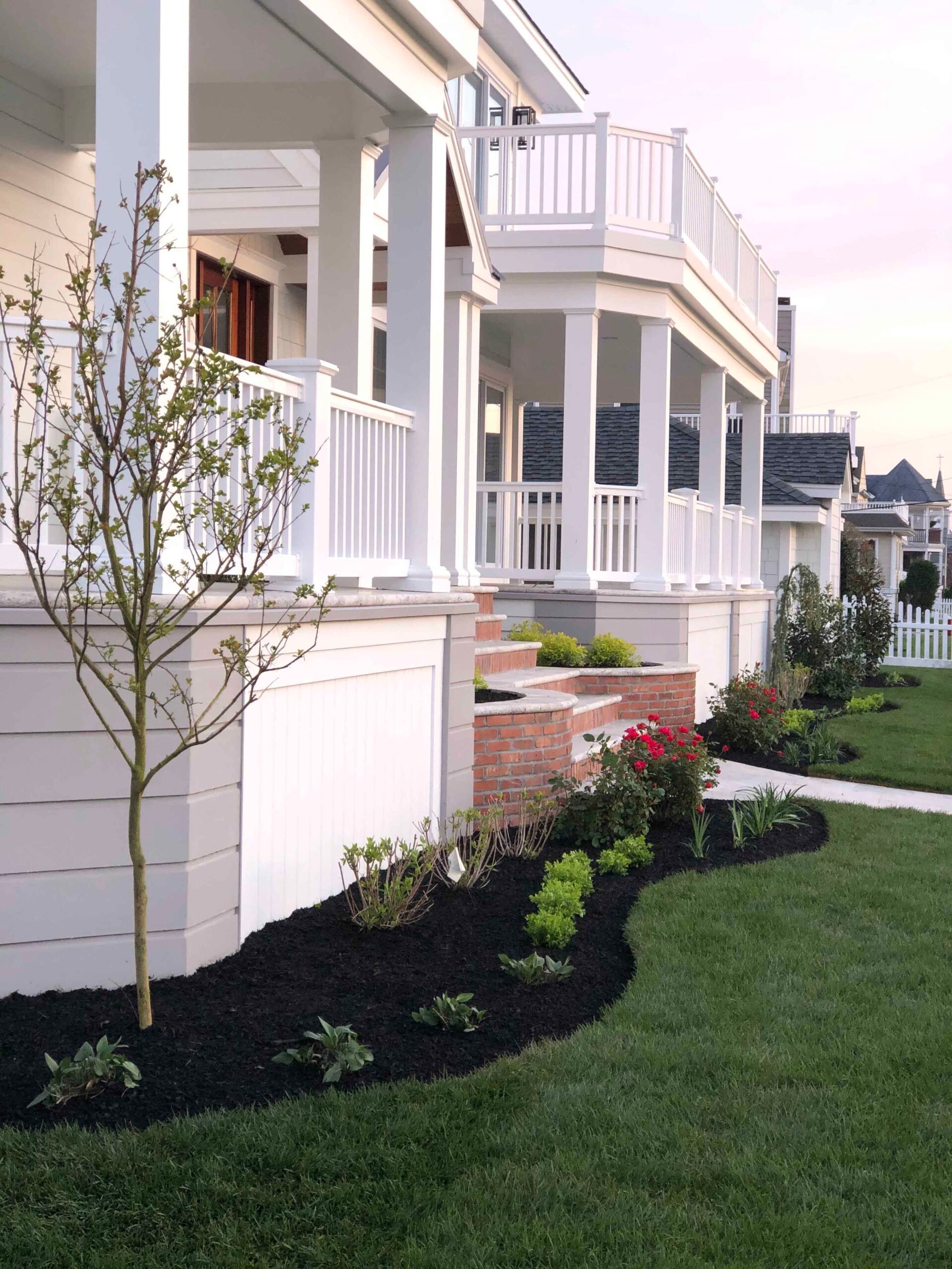 A curved front foundation planting bed along a coastal-style white house features newly installed shrubs, groundcover, and a young deciduous tree planted in dark black mulch with a clean curved edge. A mowed green lawn borders the bed, and brick porch step