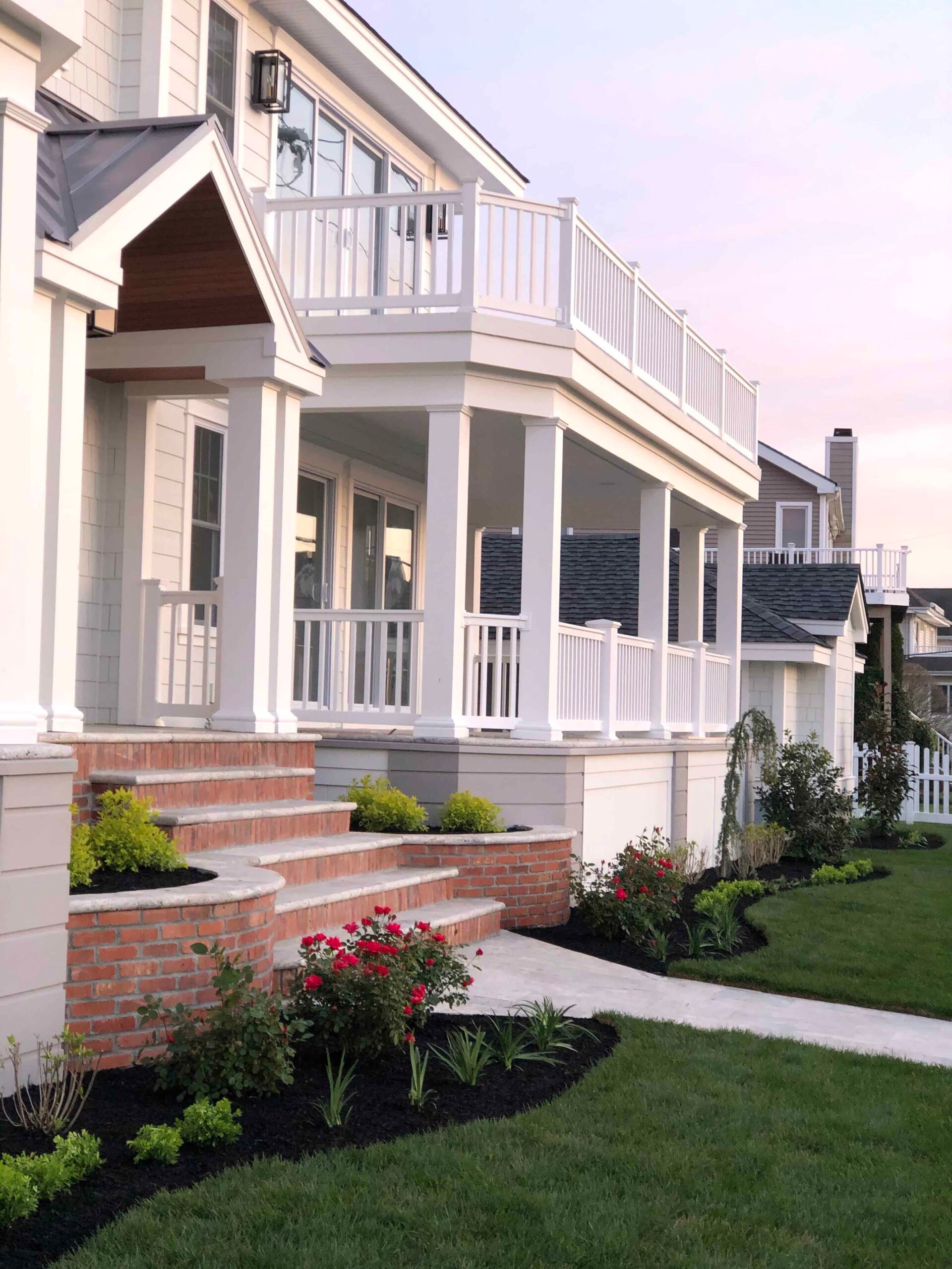 Freshly mulched black planting beds line a white colonial home's front entry, featuring red knockout roses, low boxwood shrubs, and ornamental grasses flanking brick steps with a concrete walkway. A manicured green lawn extends along the property.