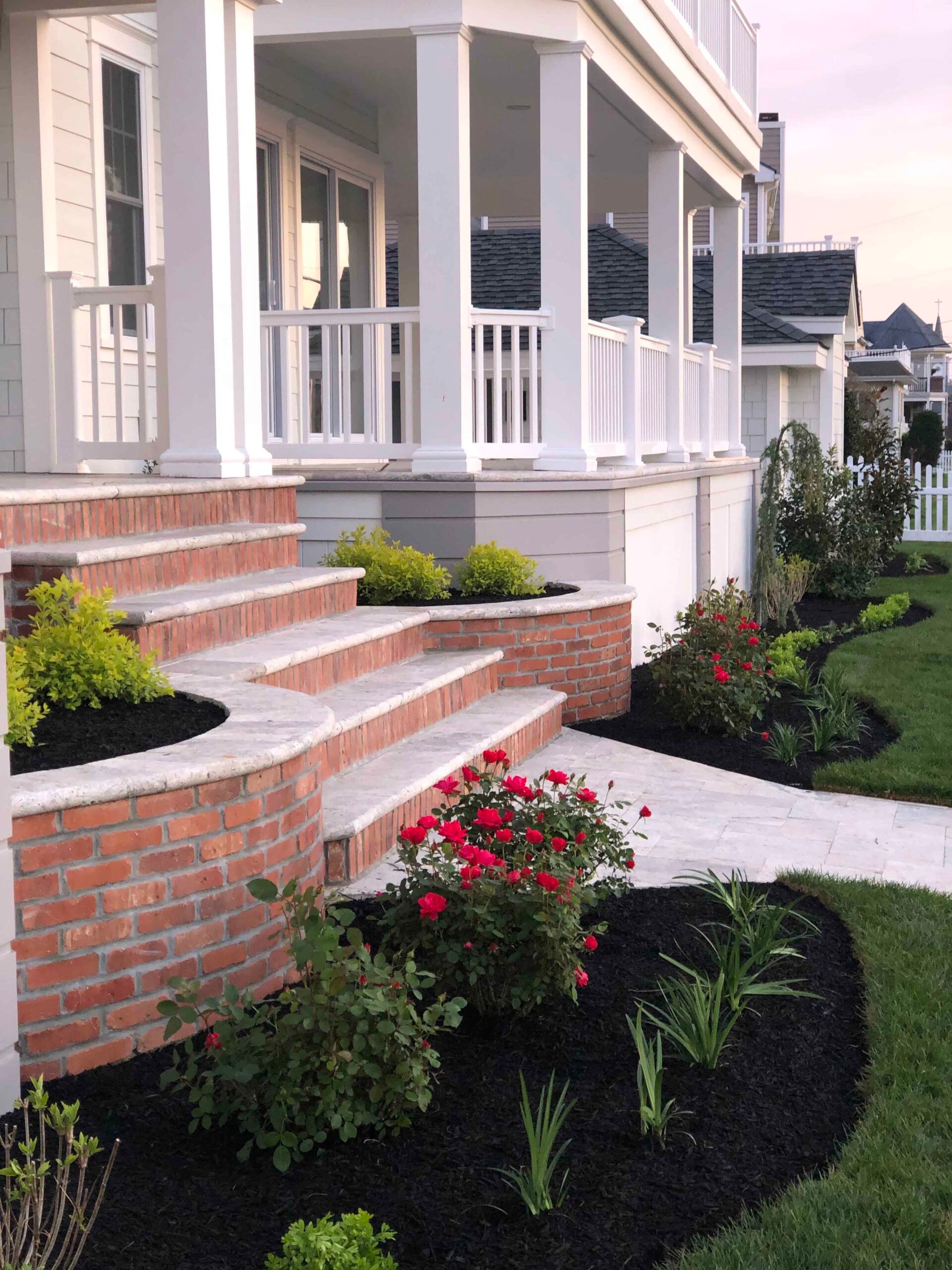 Curved brick steps with a bluestone or granite cap lead up to a covered front porch, flanked by planting beds filled with black mulch containing red knockout roses, ornamental grasses, low shrubs, and ground cover. A concrete paver walkway borders the beds