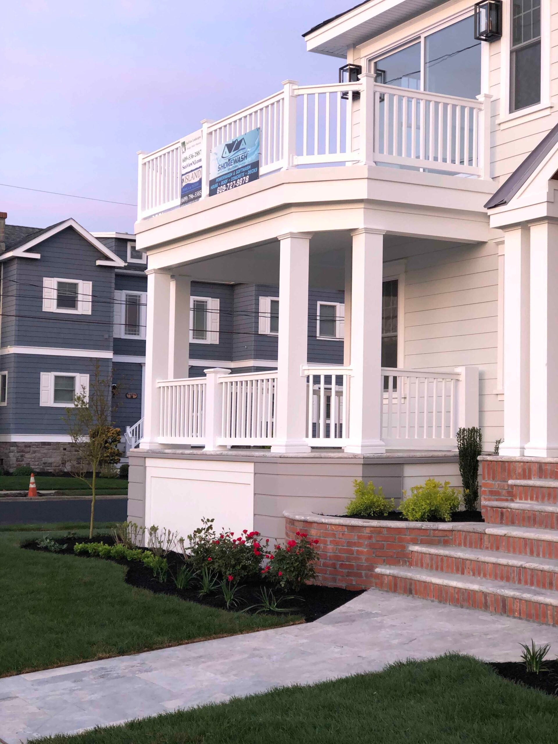 A new construction home features freshly laid turf flanking a concrete walkway, with black-mulched planting beds containing red roses and low shrubs along brick porch steps under an evening sky.