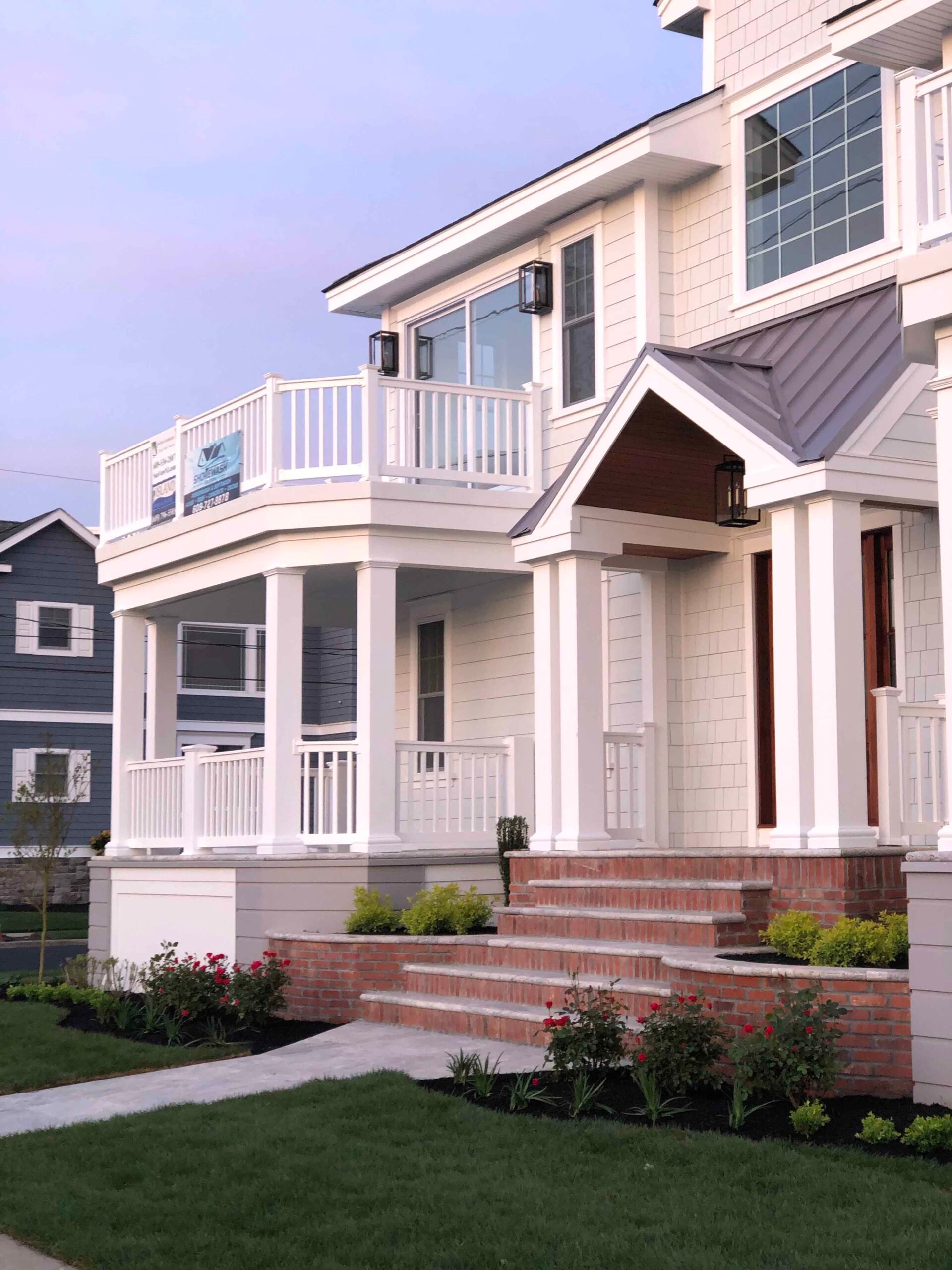 A coastal home's front facade shows brick entry steps flanked by foundation planting beds containing low shrubs and small red-flowering plants set in dark mulch, with a freshly mowed green lawn in the foreground and a covered front porch above.