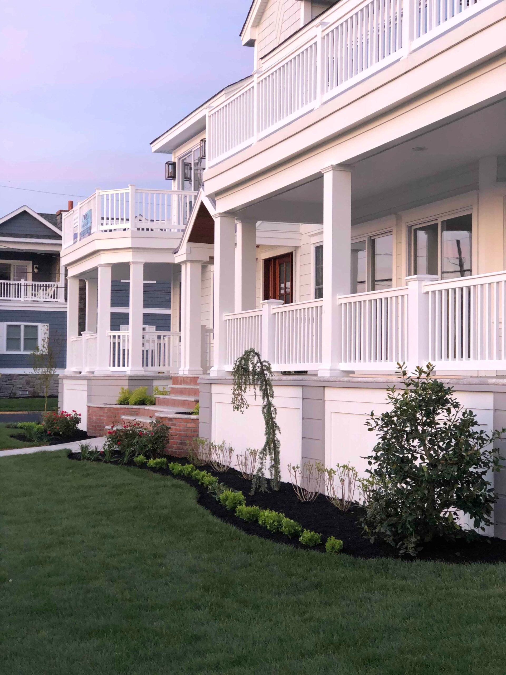 A freshly installed front foundation planting bed runs along the base of a white coastal-style home with covered porches. The bed contains mixed shrubs including a weeping specimen and broadleaf evergreens planted in dark black mulch with a defined curved 
