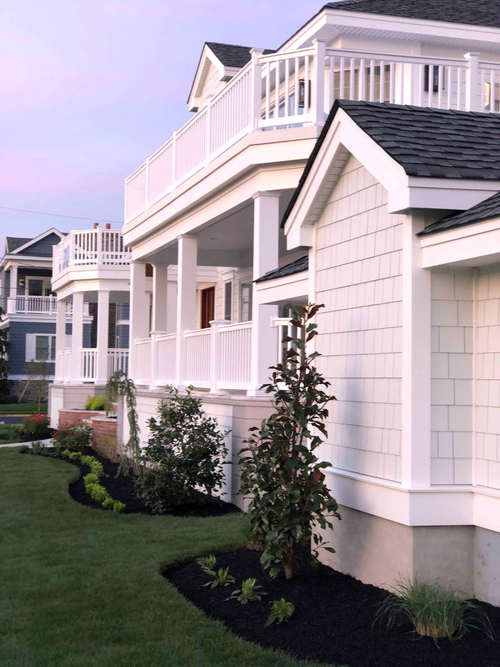 Curved planting beds along a multi-story coastal home's foundation are filled with dark black mulch, young ornamental shrubs including a columnar tree resembling a serviceberry or magnolia, and low spreading groundcover plants. The lawn is freshly mowed an