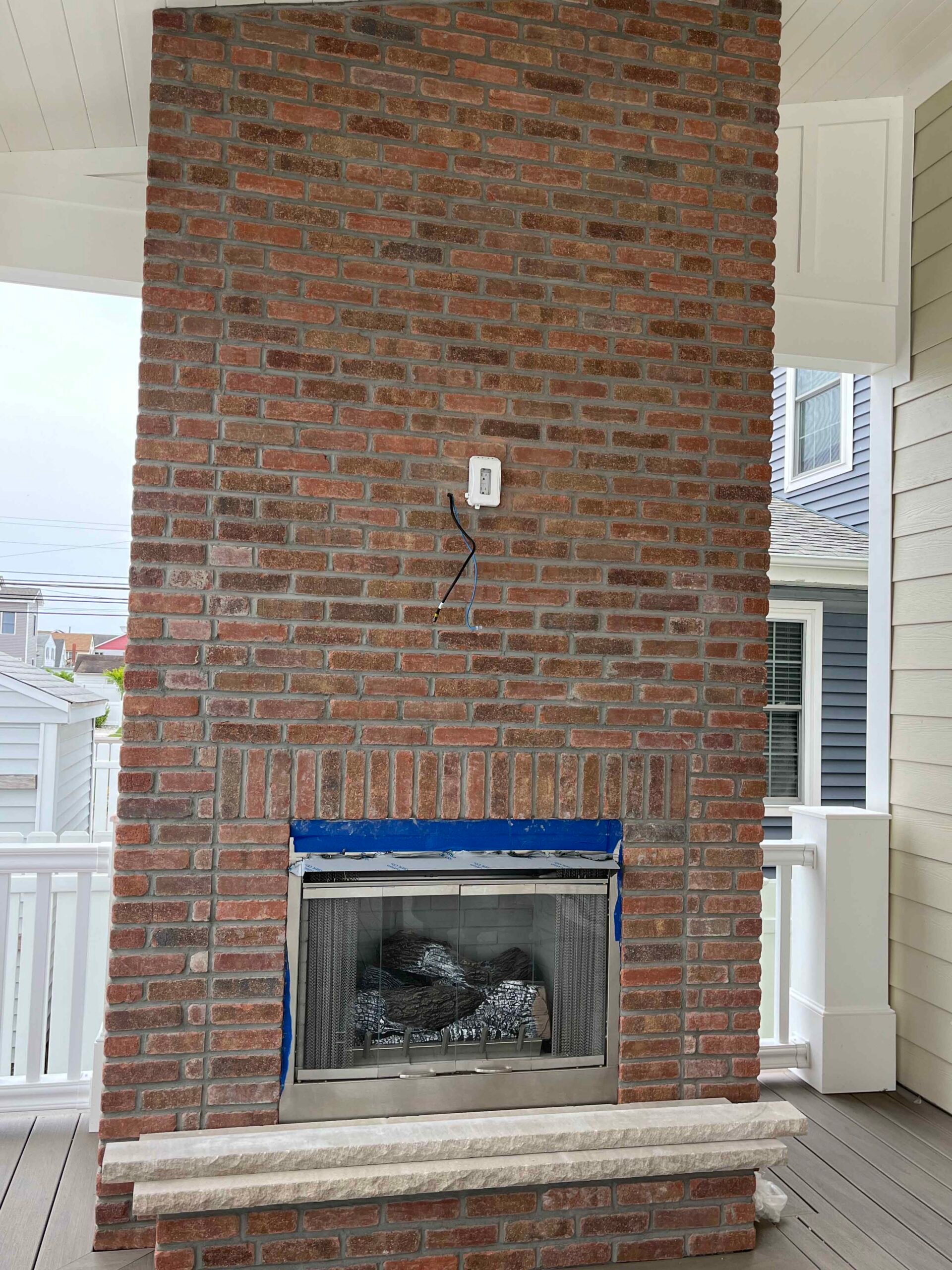 A full-height red brick outdoor fireplace with a stainless steel firebox insert sits on a covered porch with composite decking. Blue painter's tape frames the firebox opening, indicating recent installation or finishing work.