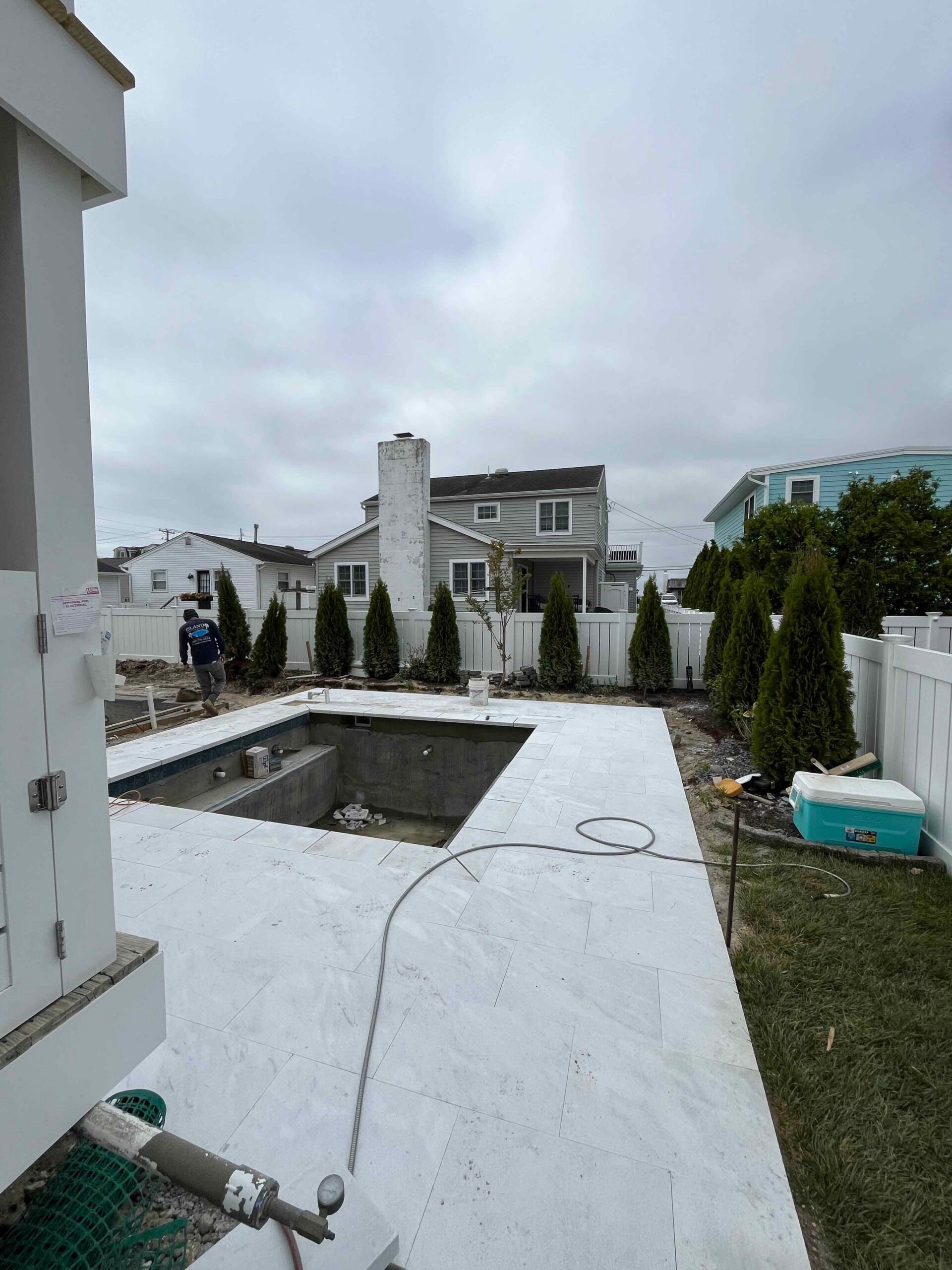 A rectangular in-ground pool surrounded by large-format light gray porcelain or marble tile decking is shown mid-installation, with construction materials visible and a row of emerald arborvitae along a white vinyl fence at the rear. This is an in-progress