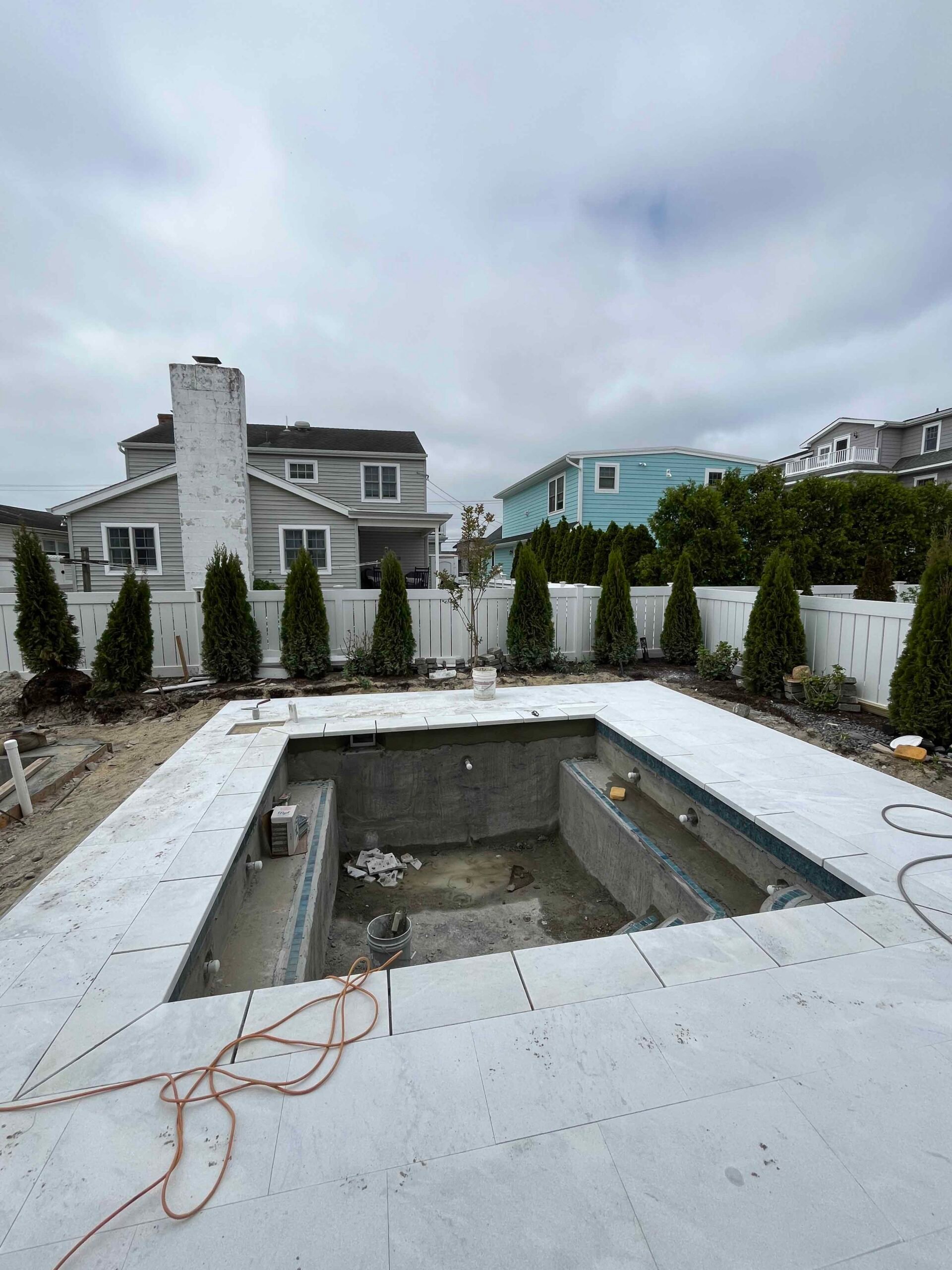 An in-ground pool under construction is shown with large-format light-gray marble or porcelain tile coping and surround installed. A row of emerald green arborvitae lines a white vinyl privacy fence along the back property line. This is an in-progress inst