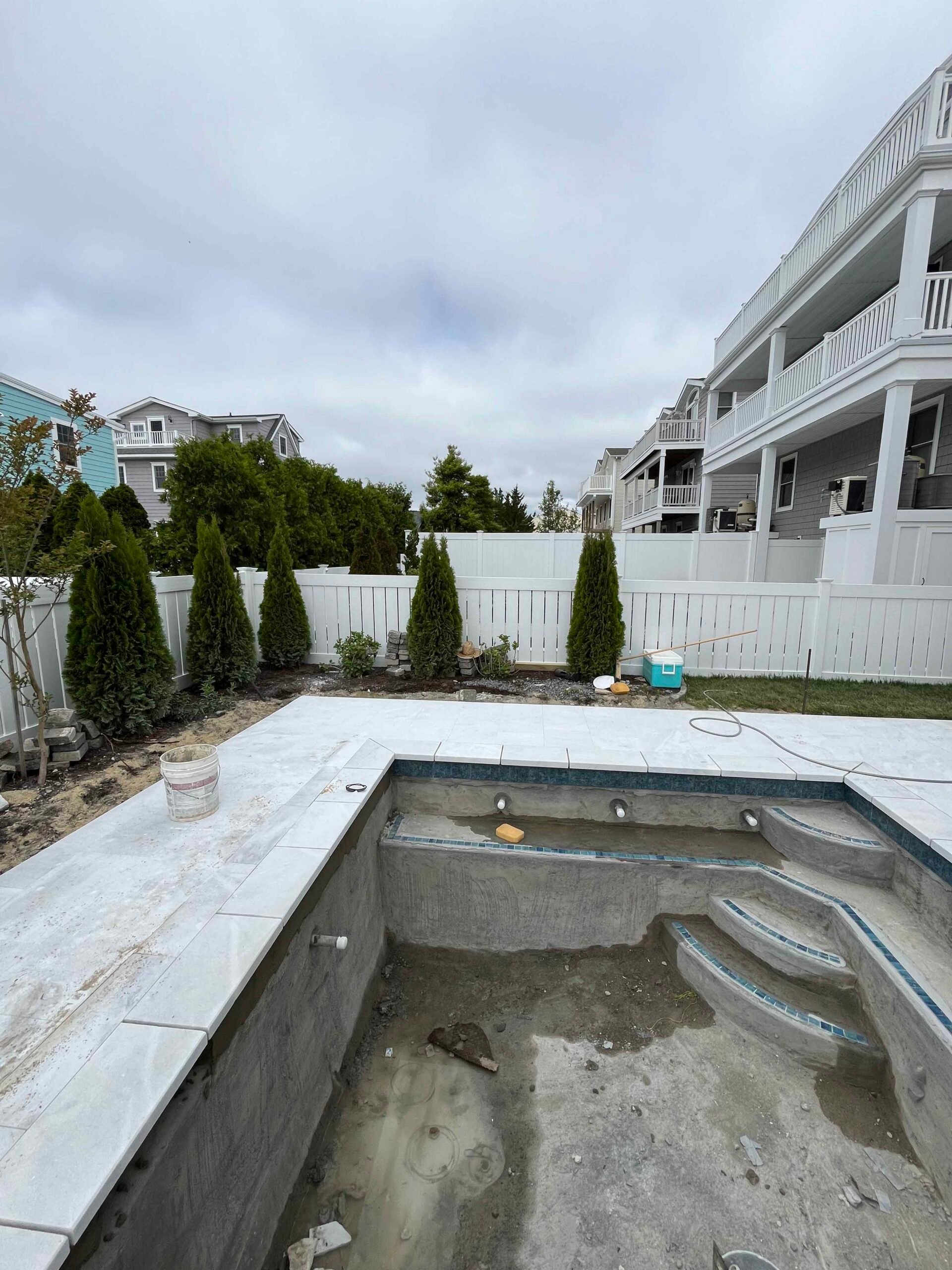An under-construction pool area showing a rectangular in-ground pool with a newly installed light-gray natural stone or large-format paver coping surround. Emerald arborvitae are planted along a white vinyl privacy fence in the background.