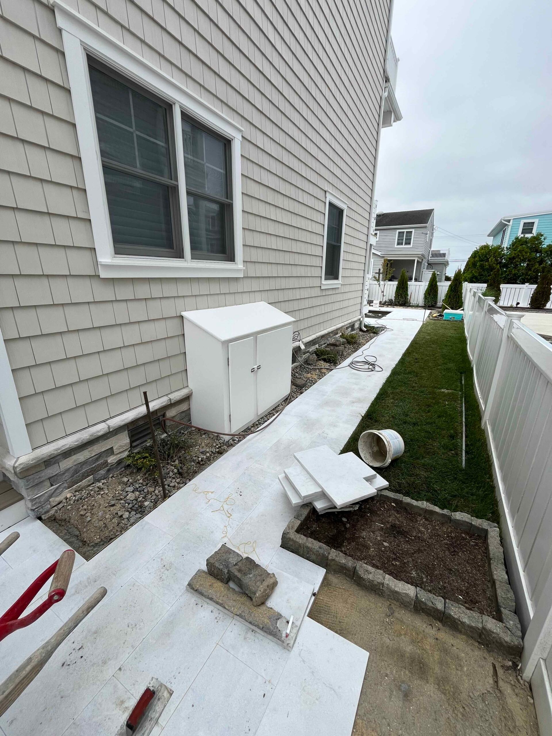 An in-progress hardscape installation showing a poured concrete walkway and patio surface along the side of a residential home. Natural stone block edging outlines a new planting bed, and materials including pavers and a concrete form tube are staged on si