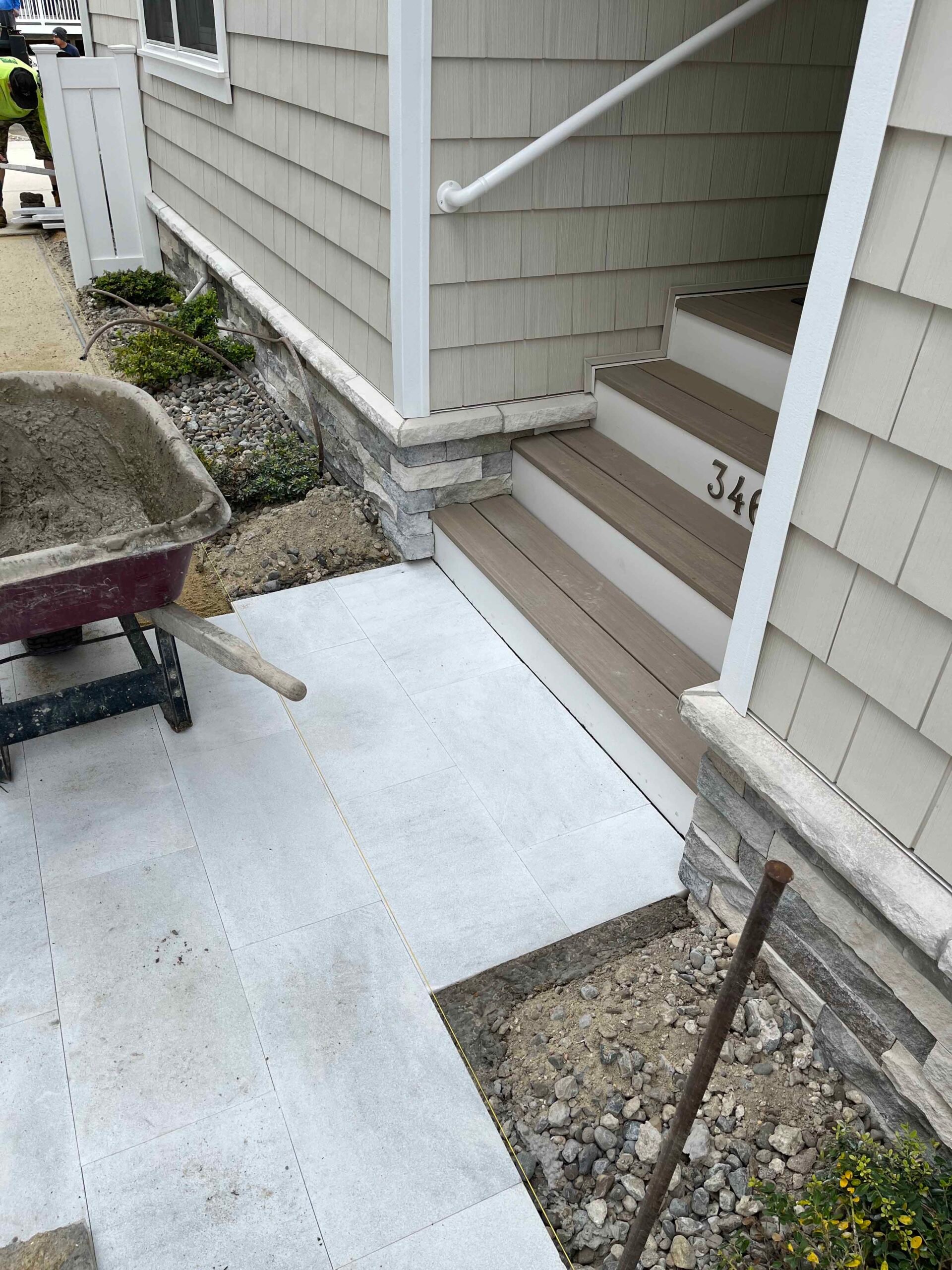 Large-format light gray porcelain or concrete pavers are being installed at a residential front entry walk mid-project, with a wheelbarrow and gravel base visible. Natural stone veneer panels clad the stair risers leading to composite decking steps at the 
