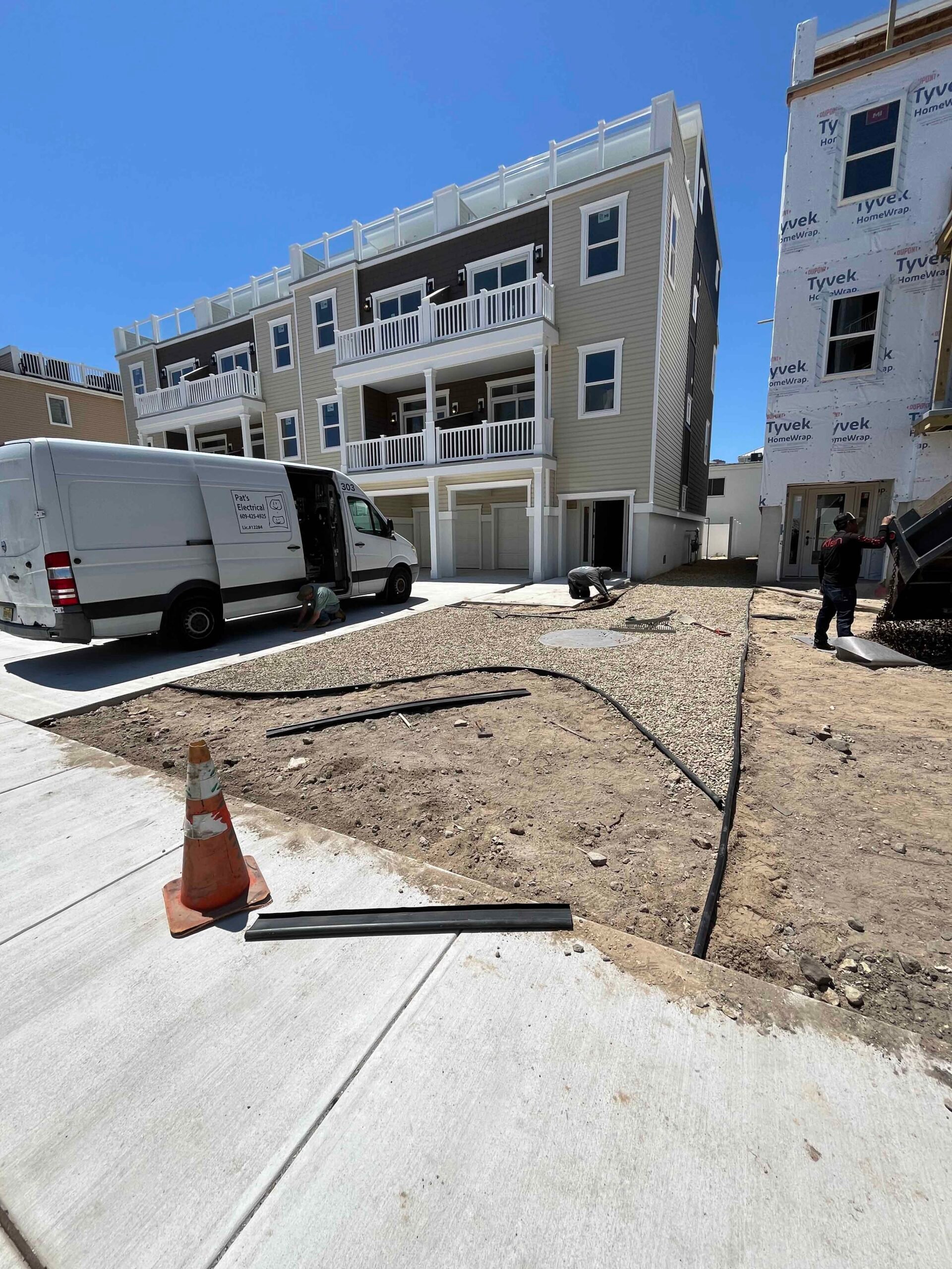 A new construction site shows a gravel base being prepared for a driveway with irrigation supply lines laid across bare soil. A concrete apron is poured at the garage entrance and workers are on site next to a multi-story residential building.