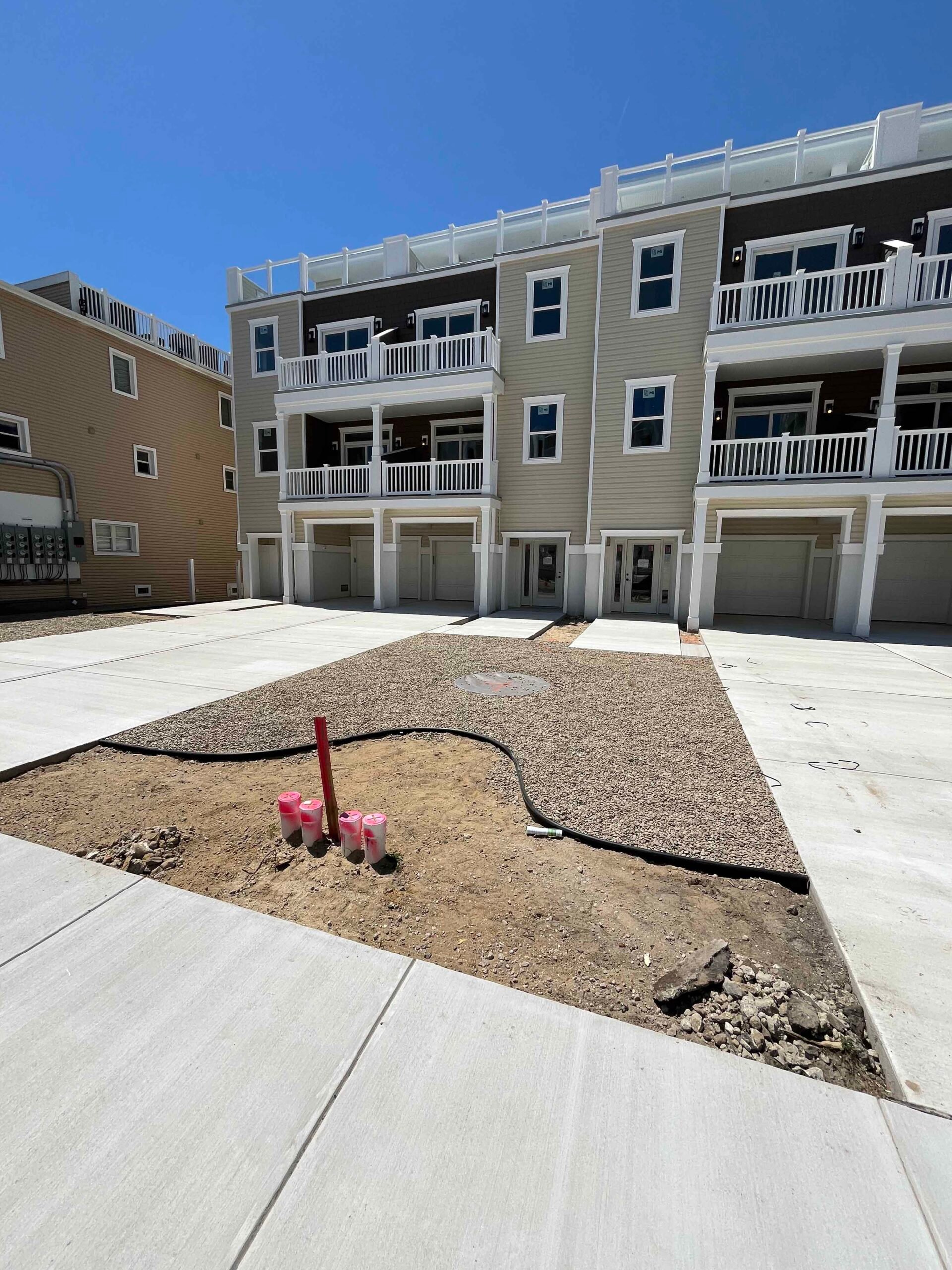 A large concrete slab driveway is newly poured in front of a multi-unit coastal townhouse, with a compacted gravel planting bed area roughed in at center and irrigation lines and marker stakes visible at grade, indicating a landscape installation in progre