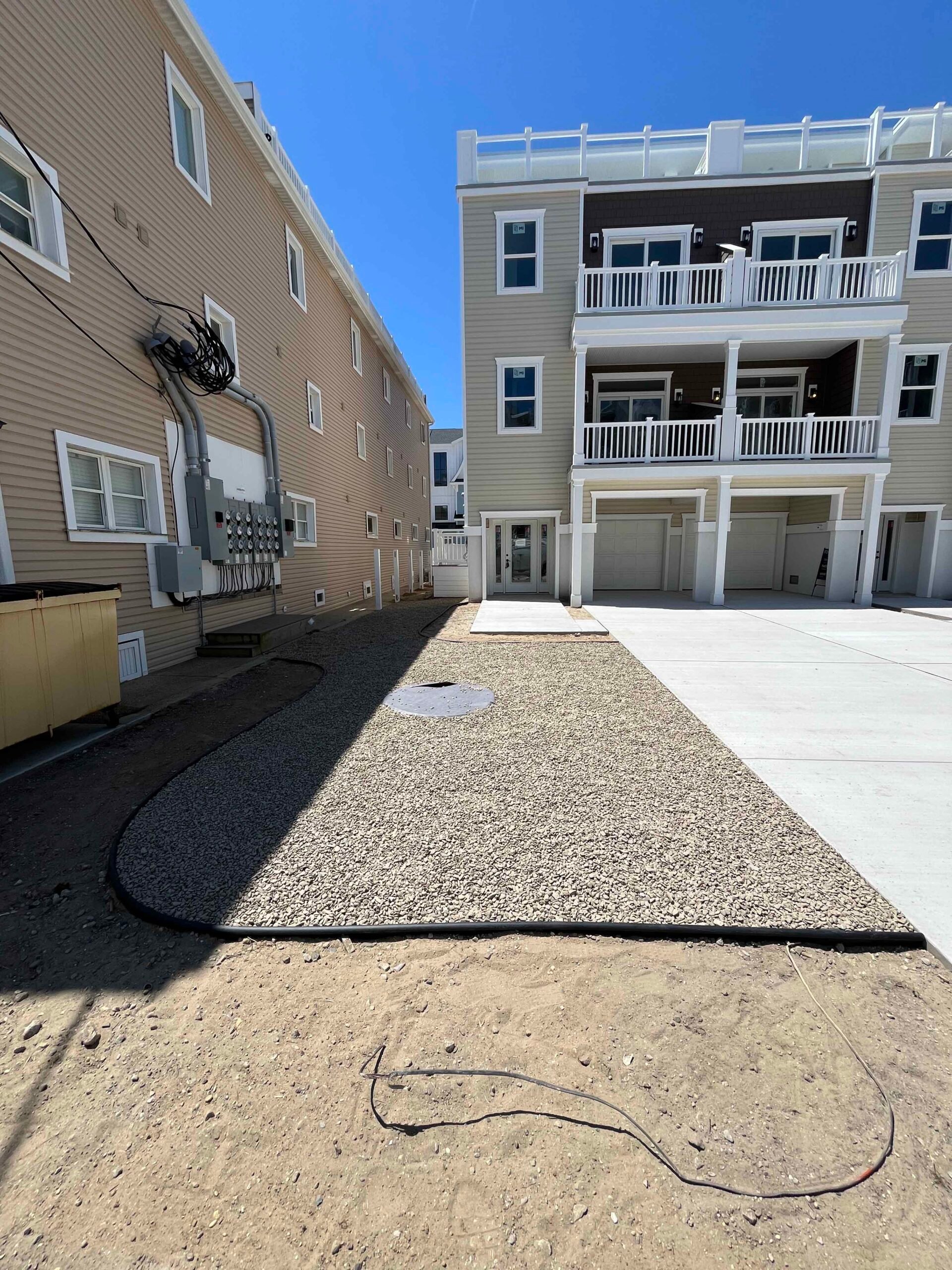 A freshly installed gravel driveway base runs between two multi-story coastal-style townhomes, with compacted crushed stone covering the full drive width. Drip irrigation lines are laid along the perimeter on bare soil, indicating the project is in progres