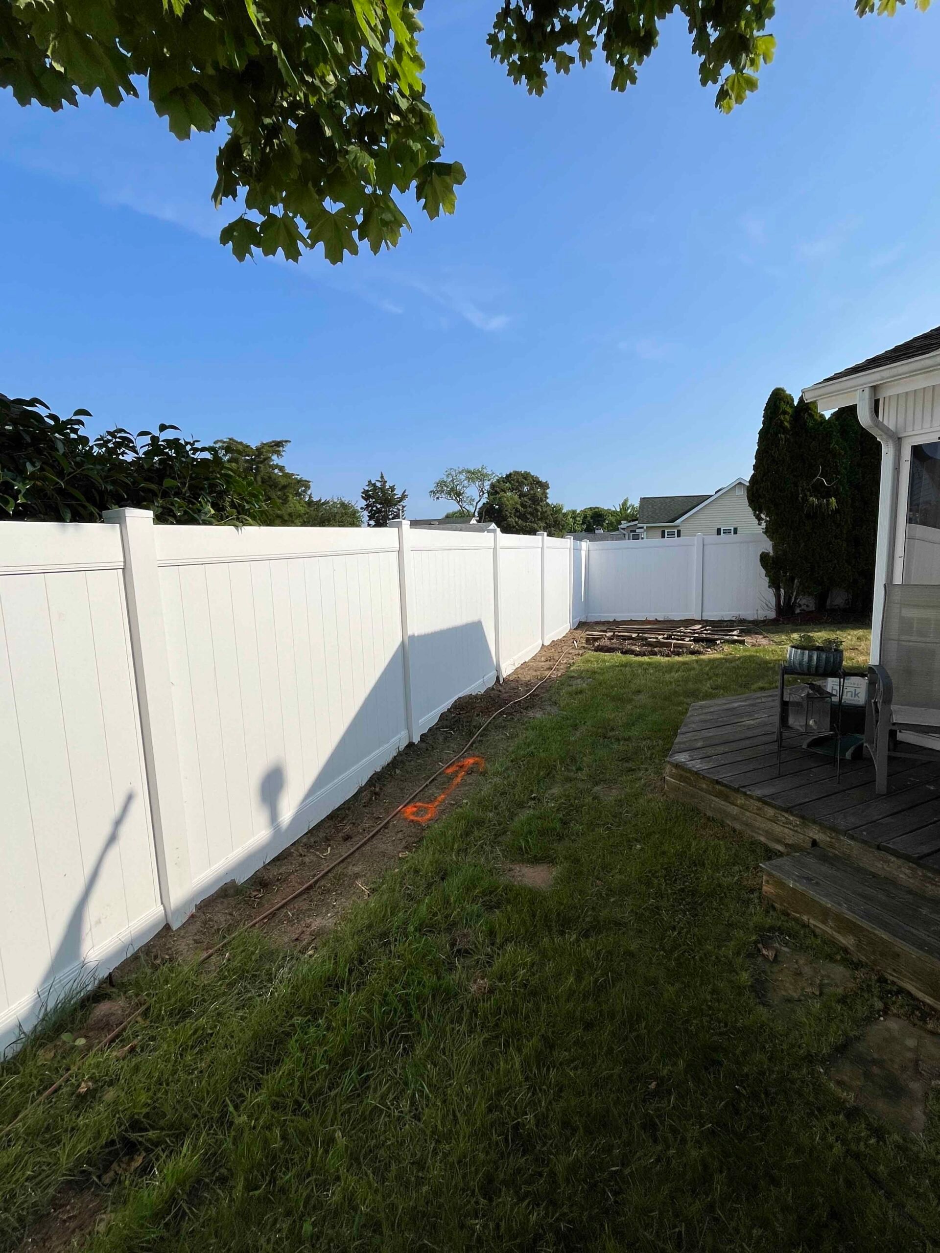 A newly installed white vinyl privacy fence lines the perimeter of a residential backyard. A narrow bare-soil planting strip runs along the fence base, and the lawn shows patchy grass with an orange irrigation or layout hose visible on the ground. A wood d
