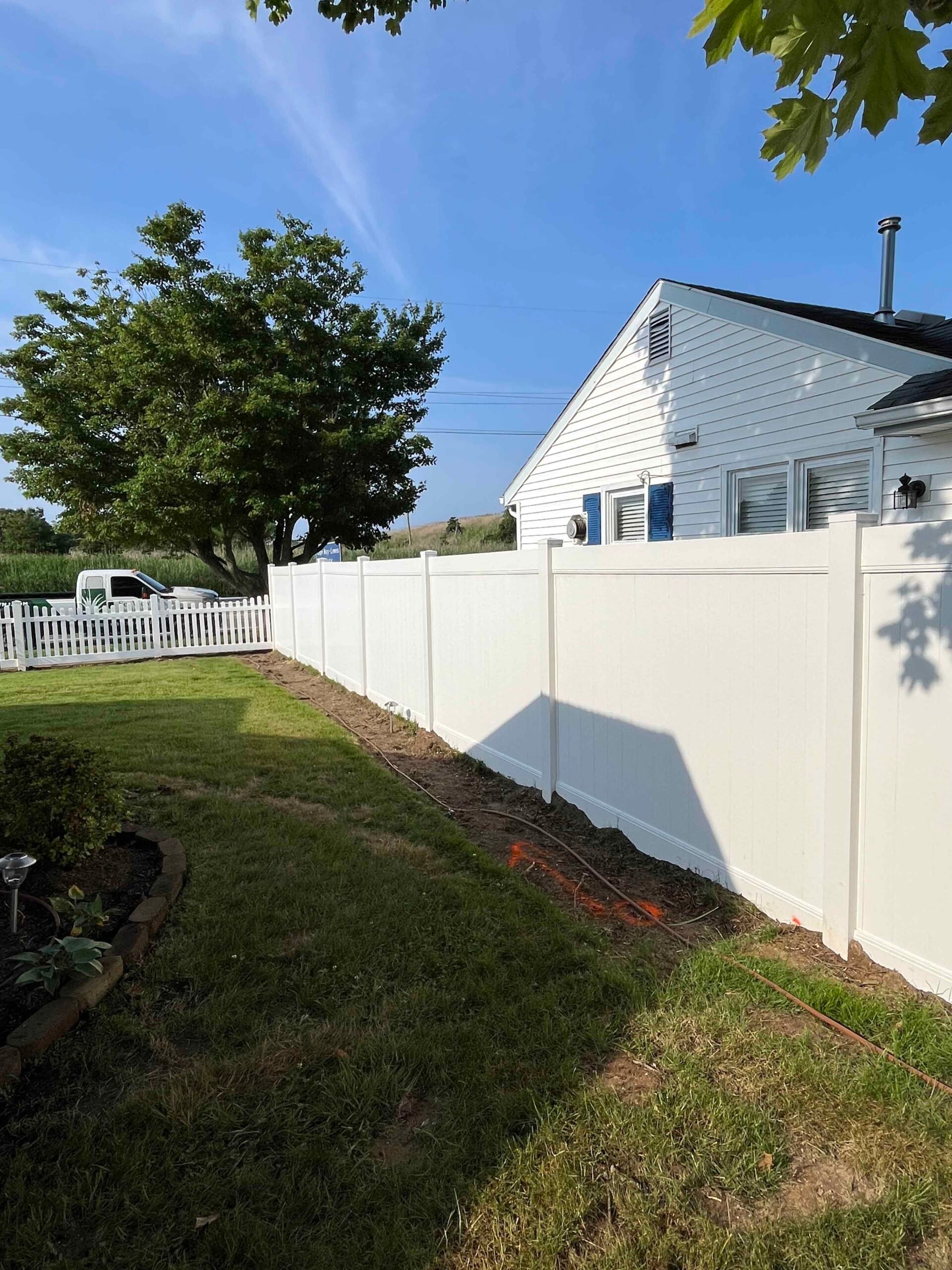 A white vinyl privacy fence runs along a residential property line beside a mowed green lawn and a mature deciduous tree. A low-voltage landscape lighting stake is visible in a mulched bed near the fence base, with sand dunes and a white picket fence visib