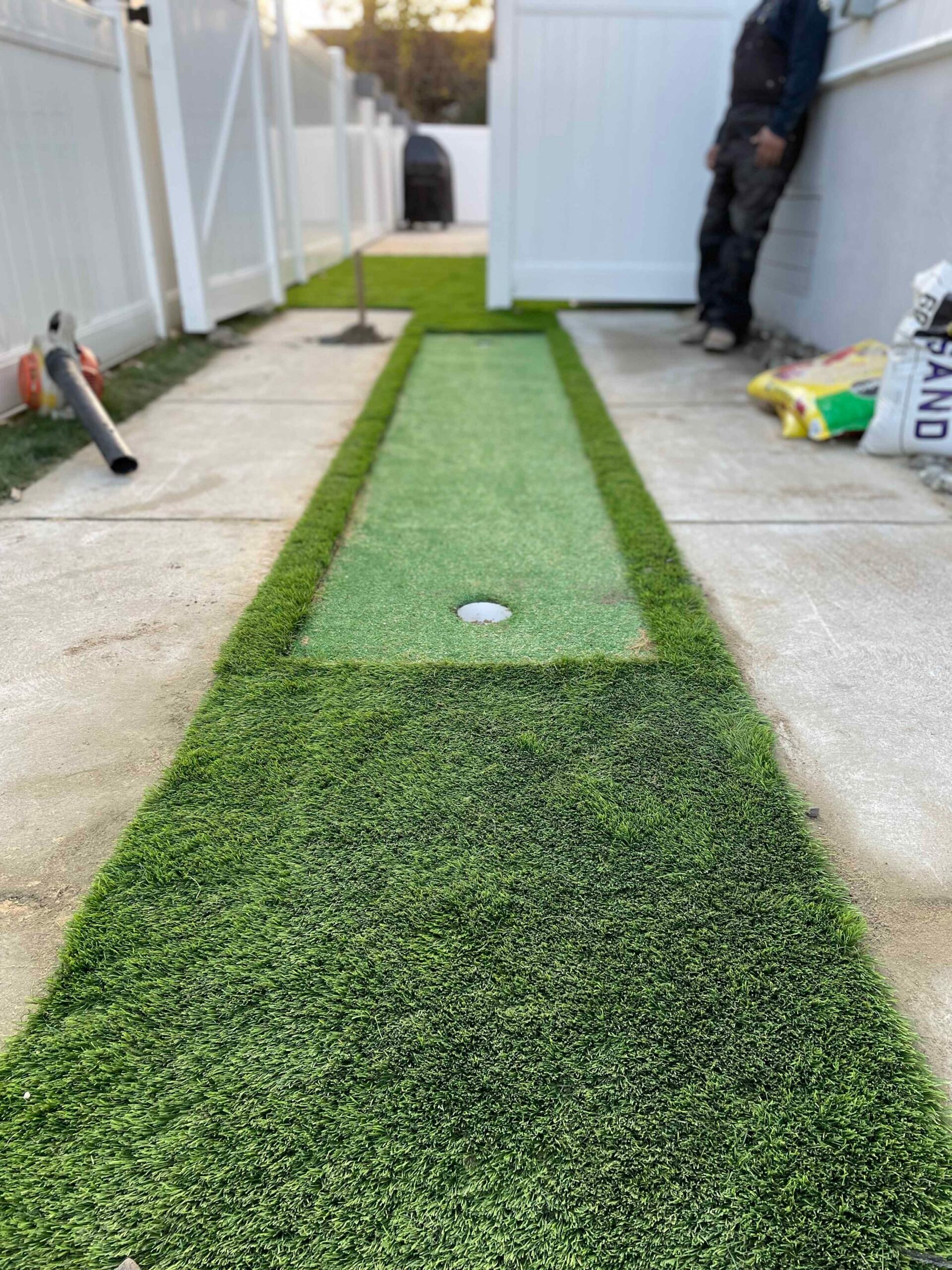 A newly installed artificial turf putting green runs the length of a narrow backyard flanked by white vinyl privacy fencing, with a single cup hole visible mid-green and concrete pavers on either side.