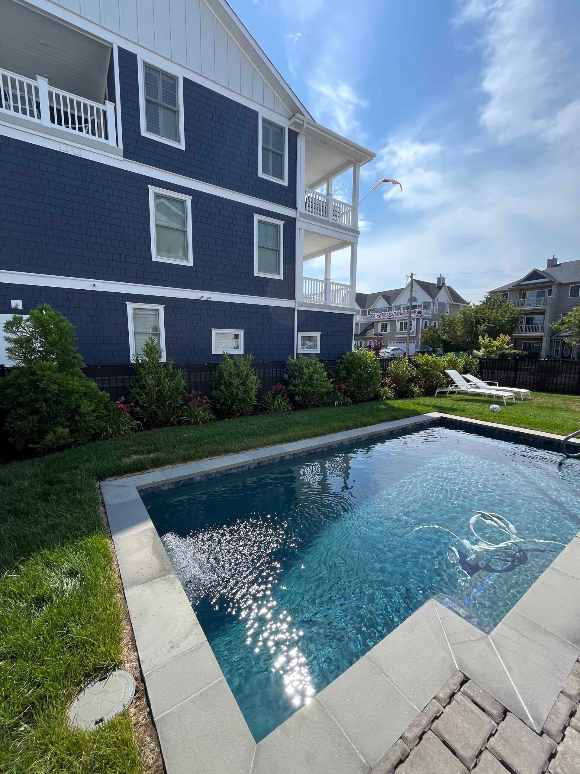 A rectangular in-ground pool is edged with bluestone or light gray paver coping and surrounded by a closely mowed lawn. A mixed shrub border running along a dark-sided multi-story house includes arborvitae, ornamental grasses, and flowering shrubs, with lo