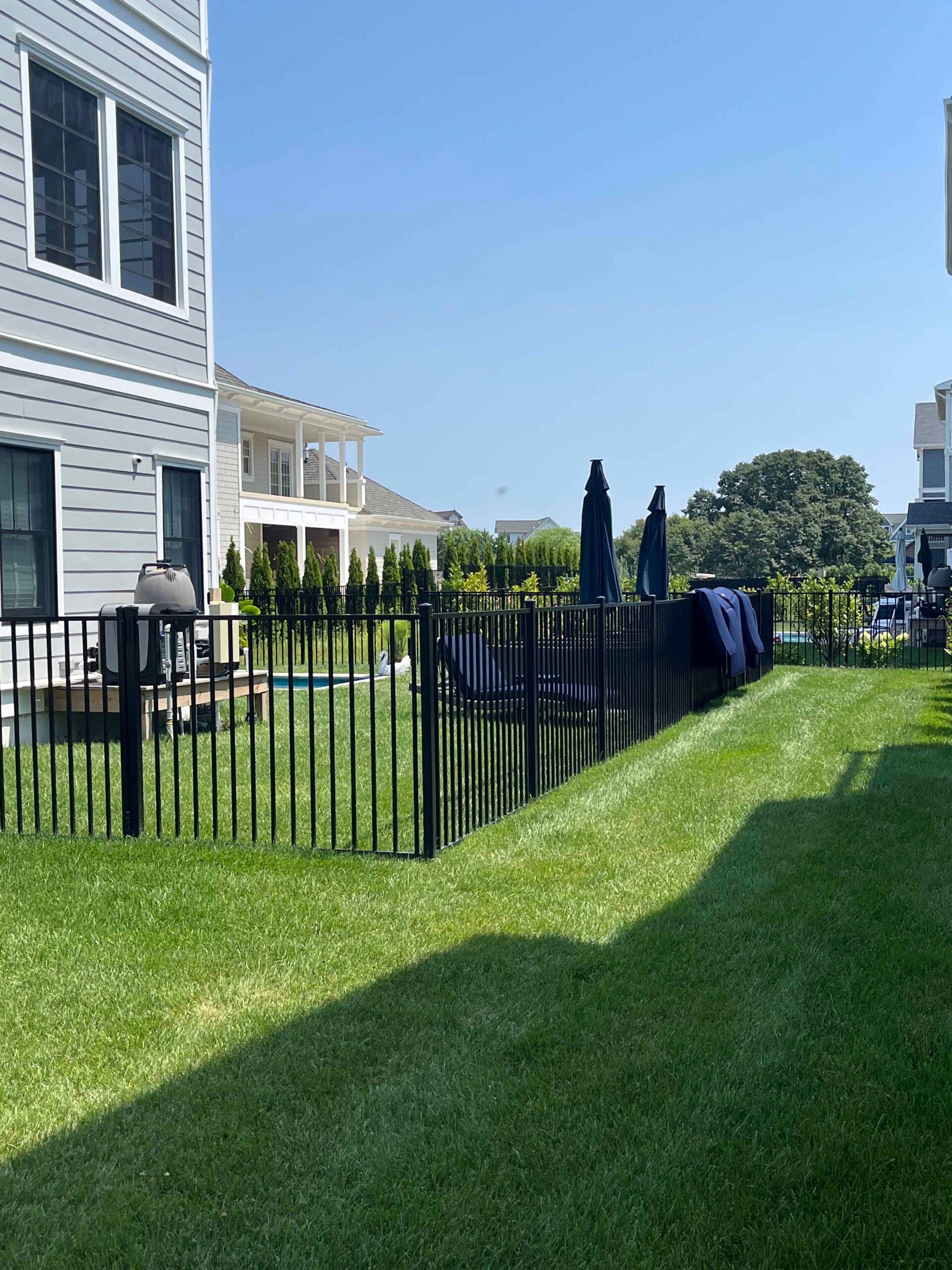 A backyard with a freshly mowed, evenly striped lawn enclosed by a black aluminum spear-top fence. Emerald green arborvitae are planted inside the fence line near a patio area with a pool and closed patio umbrellas visible in the background.