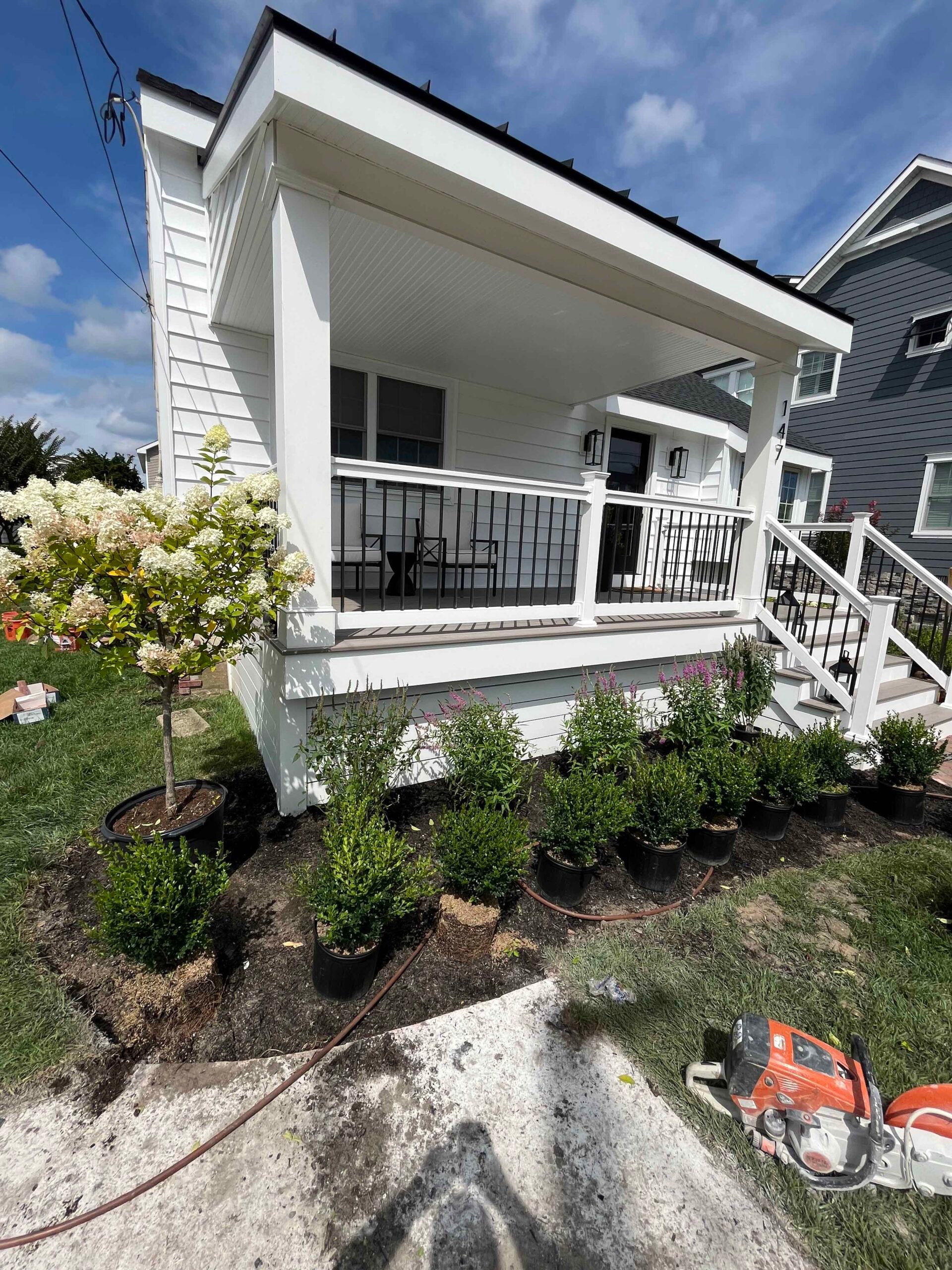 Containerized boxwood shrubs and a standard-form hydrangea tree are staged along a house foundation bed in front of a covered porch during a planting installation, with soil prepared and a garden hose visible in the foreground.
