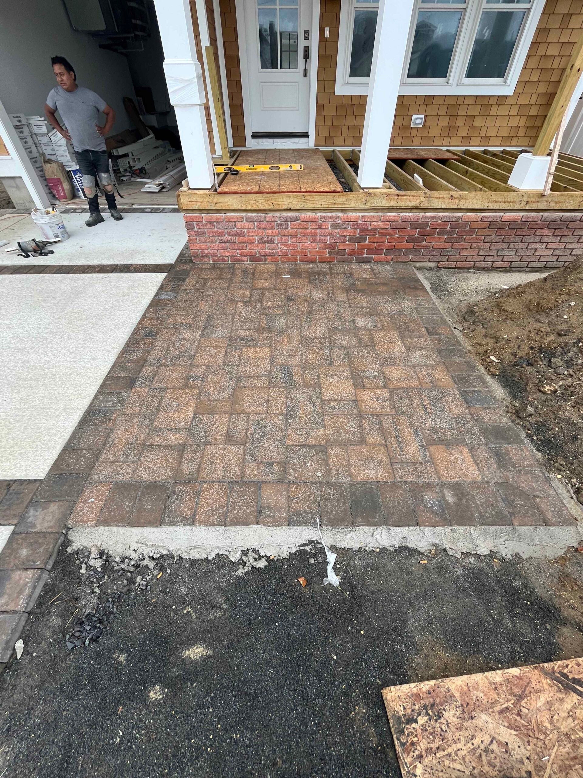 A newly laid tumbled concrete paver walkway in mixed brown and tan tones leads from a driveway to a front entry porch with a red brick skirt. The surrounding area shows exposed soil and an unfinished deck frame, indicating an active hardscape installation 