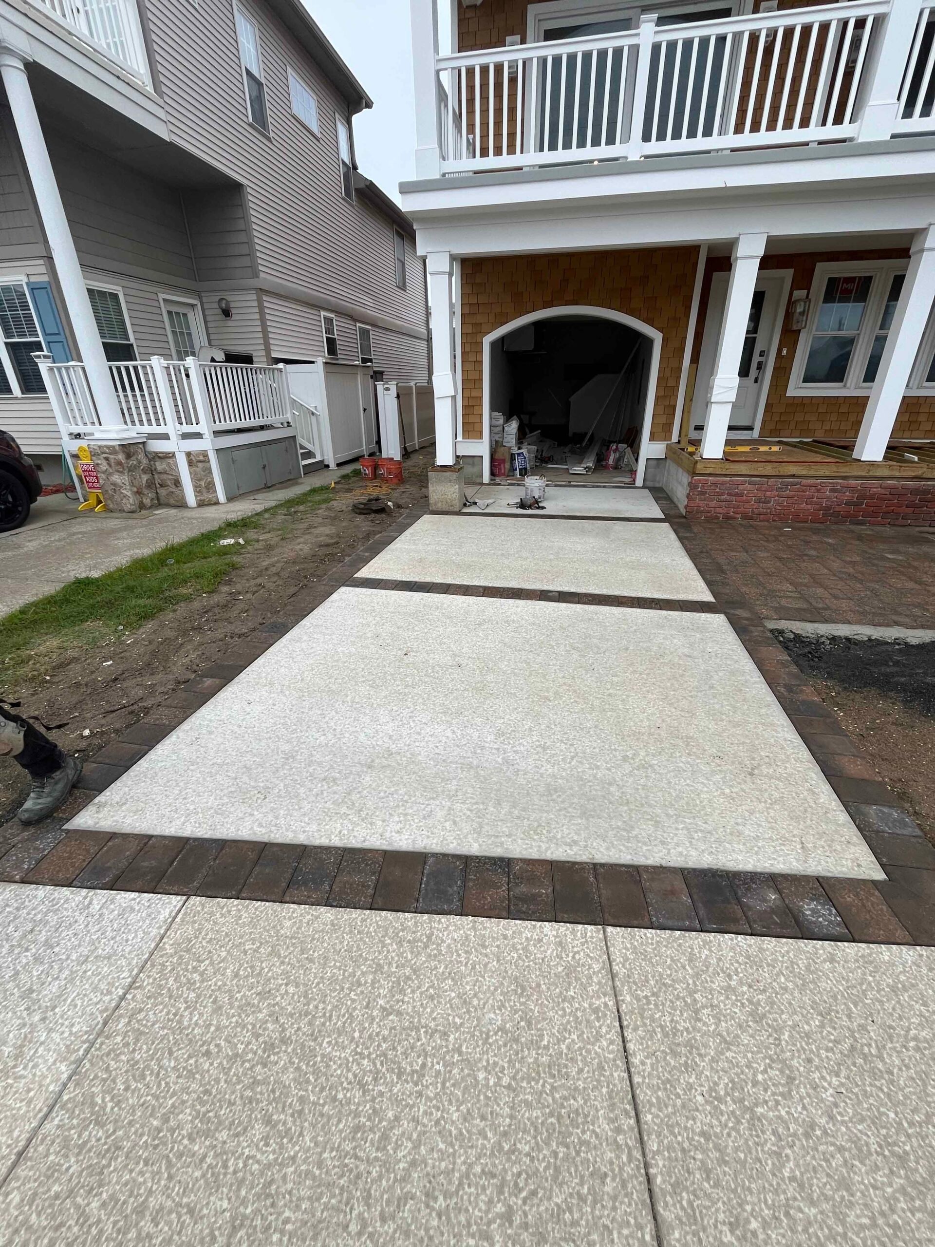 A newly installed driveway surface combines a light aggregate-finish concrete field with dark brown brick paver border strips framing the perimeter and a center band. The surrounding ground is bare soil, indicating an active construction phase.