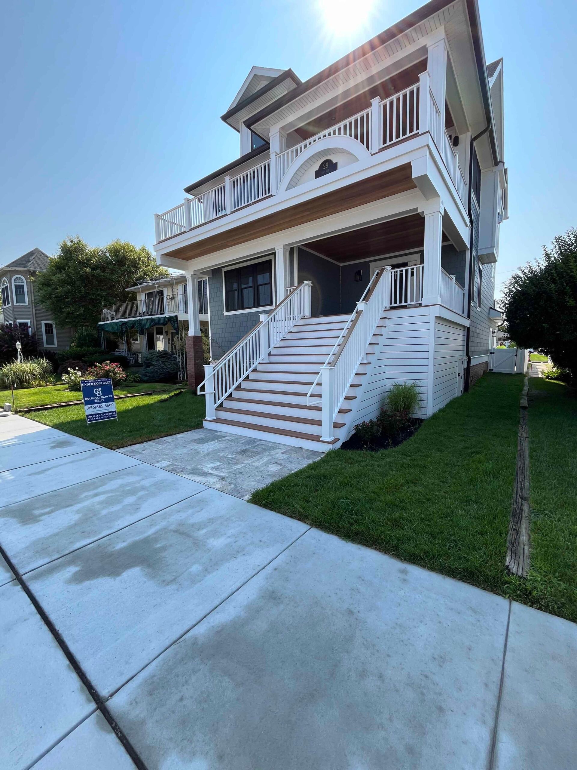 A freshly mowed front lawn borders a concrete sidewalk leading to a multi-story coastal home with white railings and a screened porch. A neatly trimmed shrub and black mulch bed are visible at the right foundation.