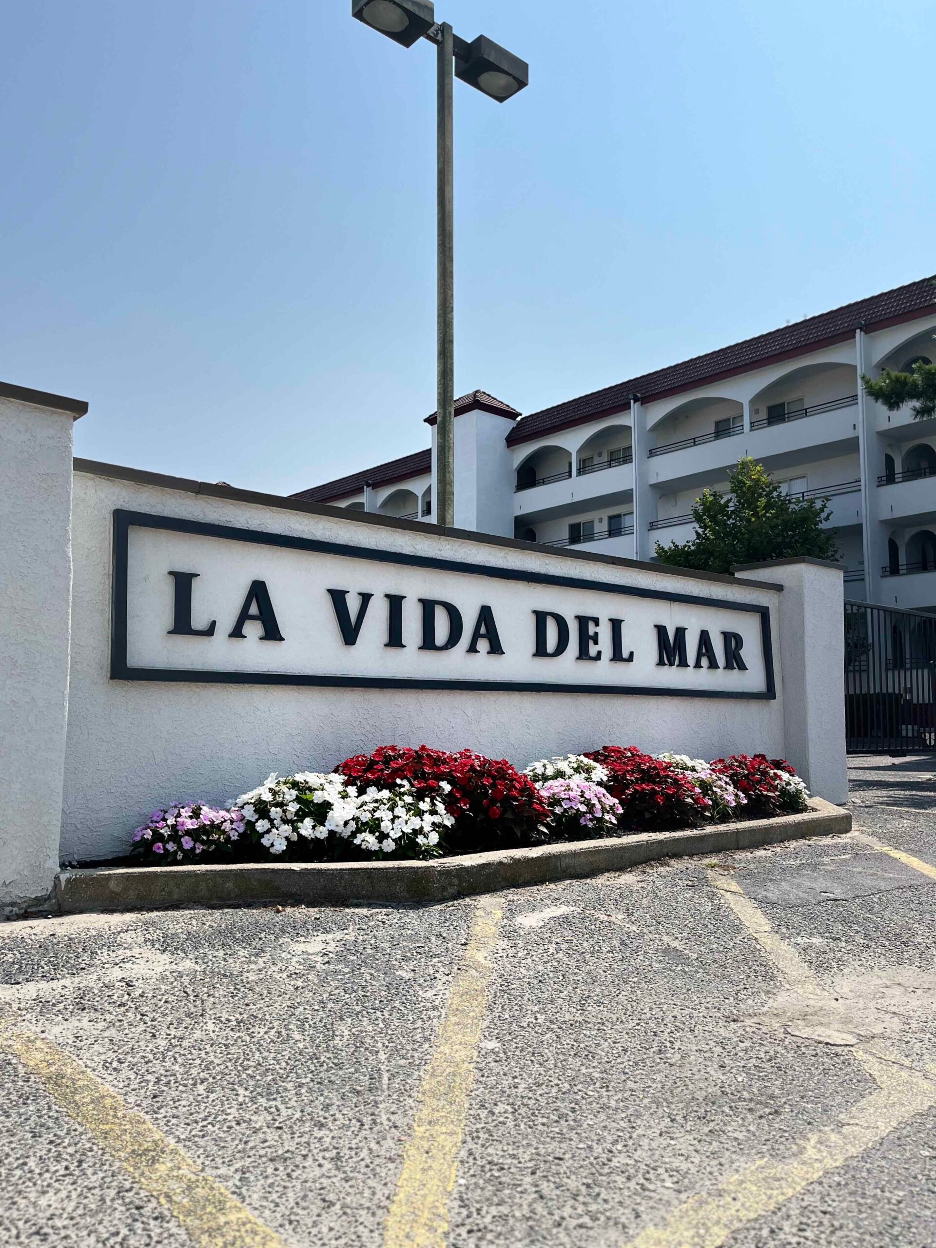 A stucco monument sign reading "La Vida Del Mar" sits in front of a commercial building with a low planting bed of red, white, and pink impatiens in full bloom at its base.