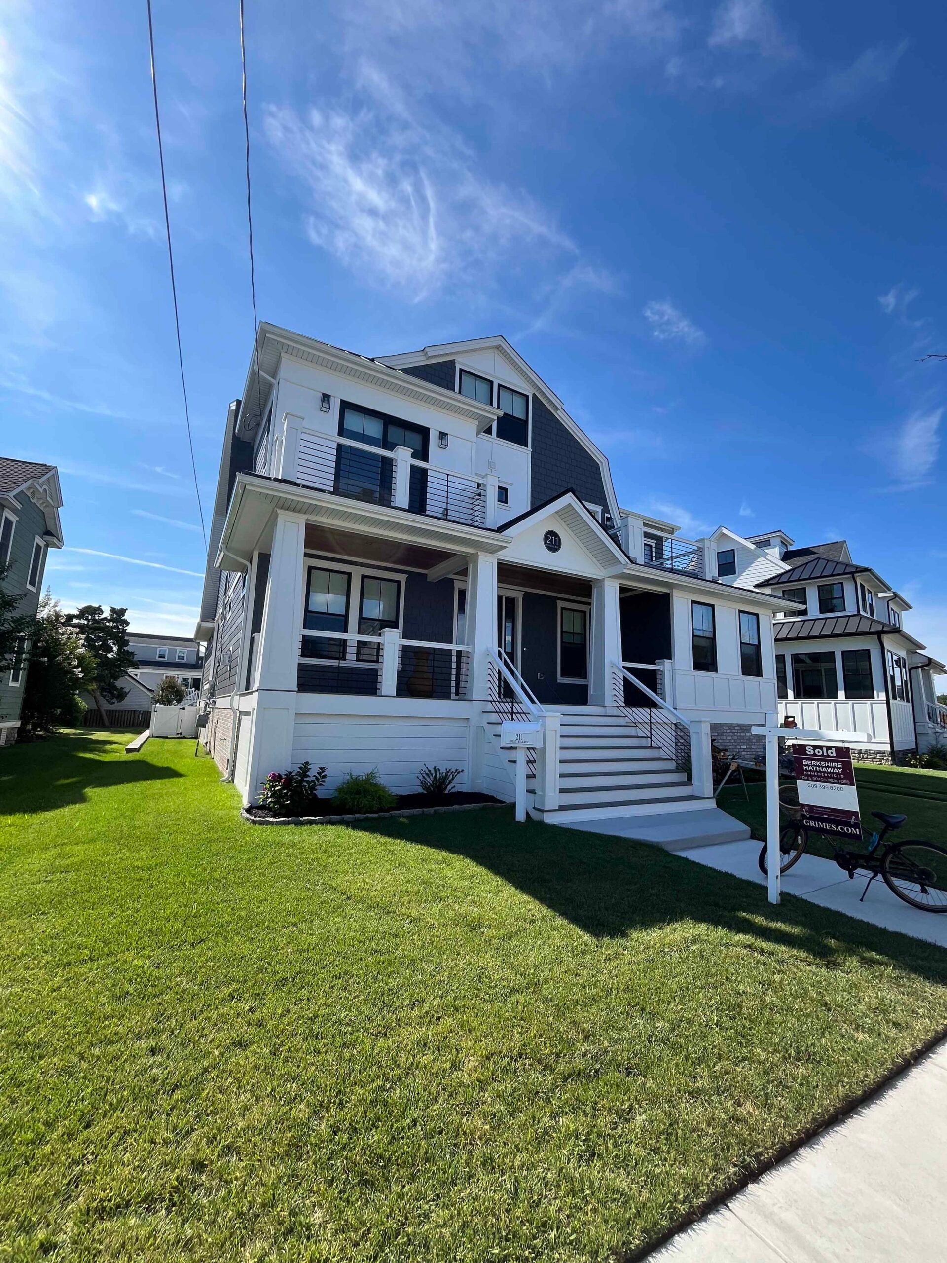 A freshly mowed, evenly striped green lawn fronts a two-story coastal-style home with white siding and dark trim. Compact low shrubs fill a narrow planting bed along the front foundation, and a concrete sidewalk runs along the right side.