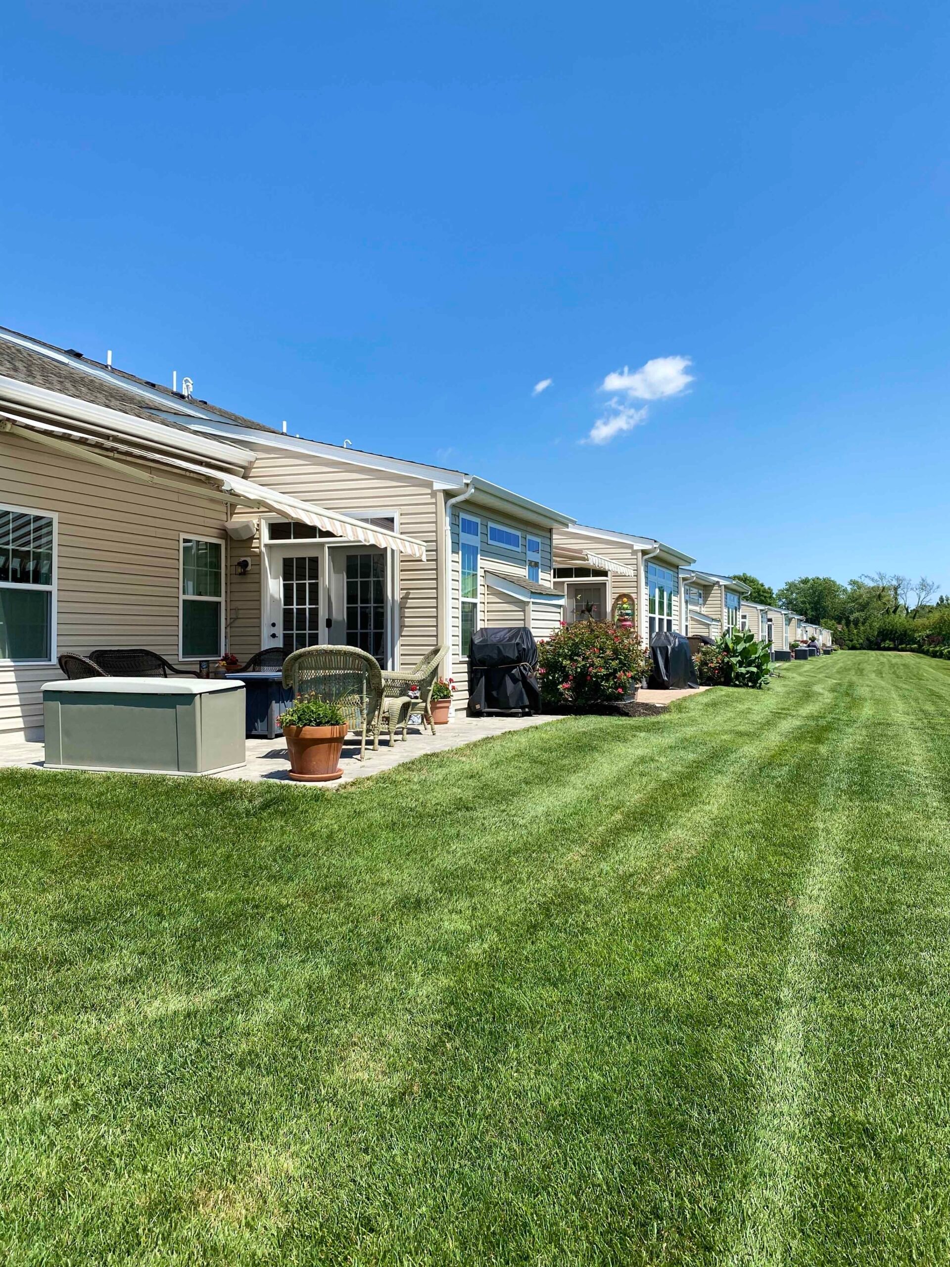 A wide expanse of freshly mowed, striped green lawn runs along the rear of a row of attached residential units with concrete patios, container plantings, and patio furniture visible along the building.