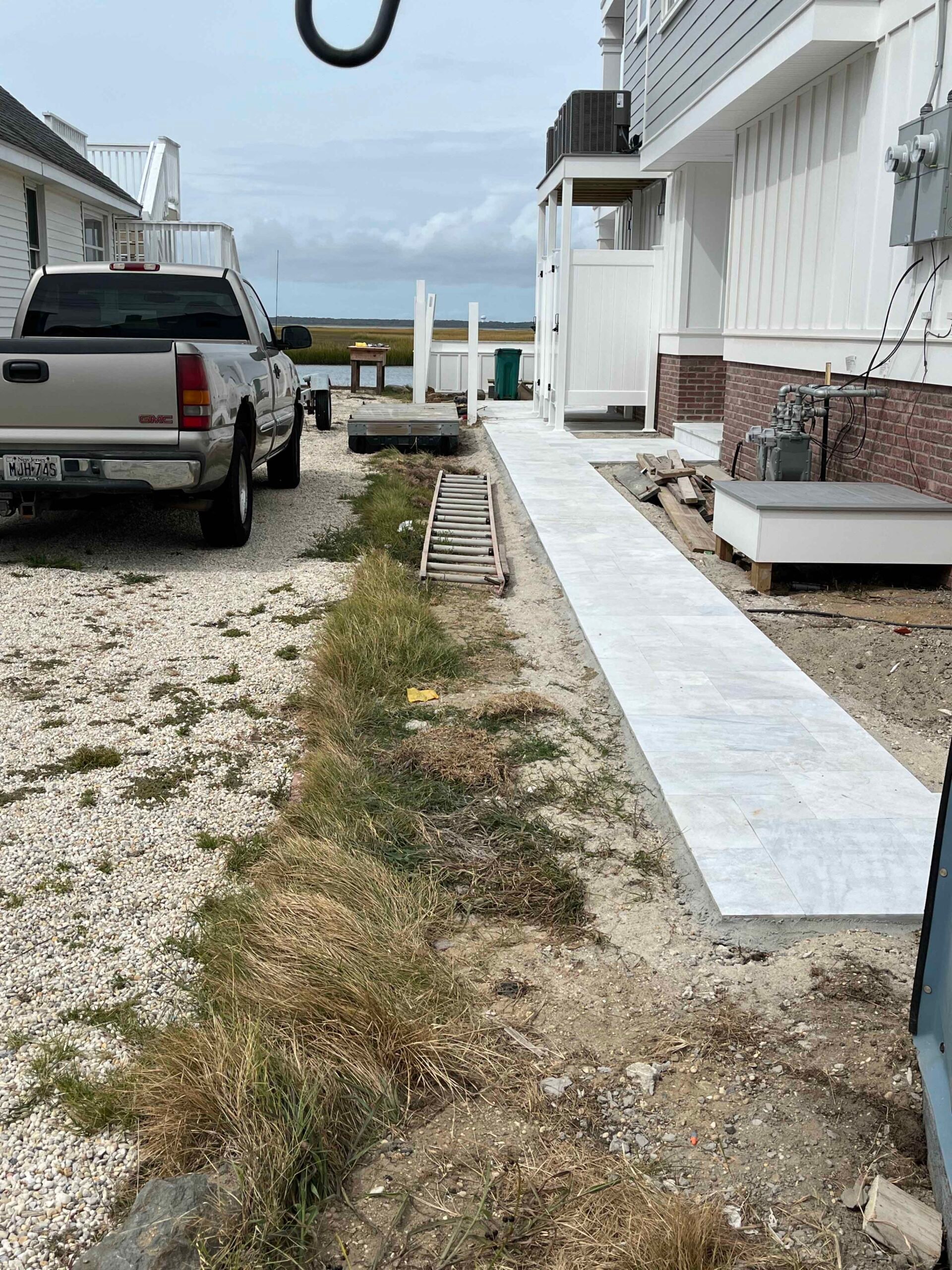 A newly installed light gray tile or large-format concrete paver walkway runs along the side of a coastal residential property. The surrounding ground is bare gravel and sandy soil with patches of beach grass. A metal drainage grate is visible mid-installa