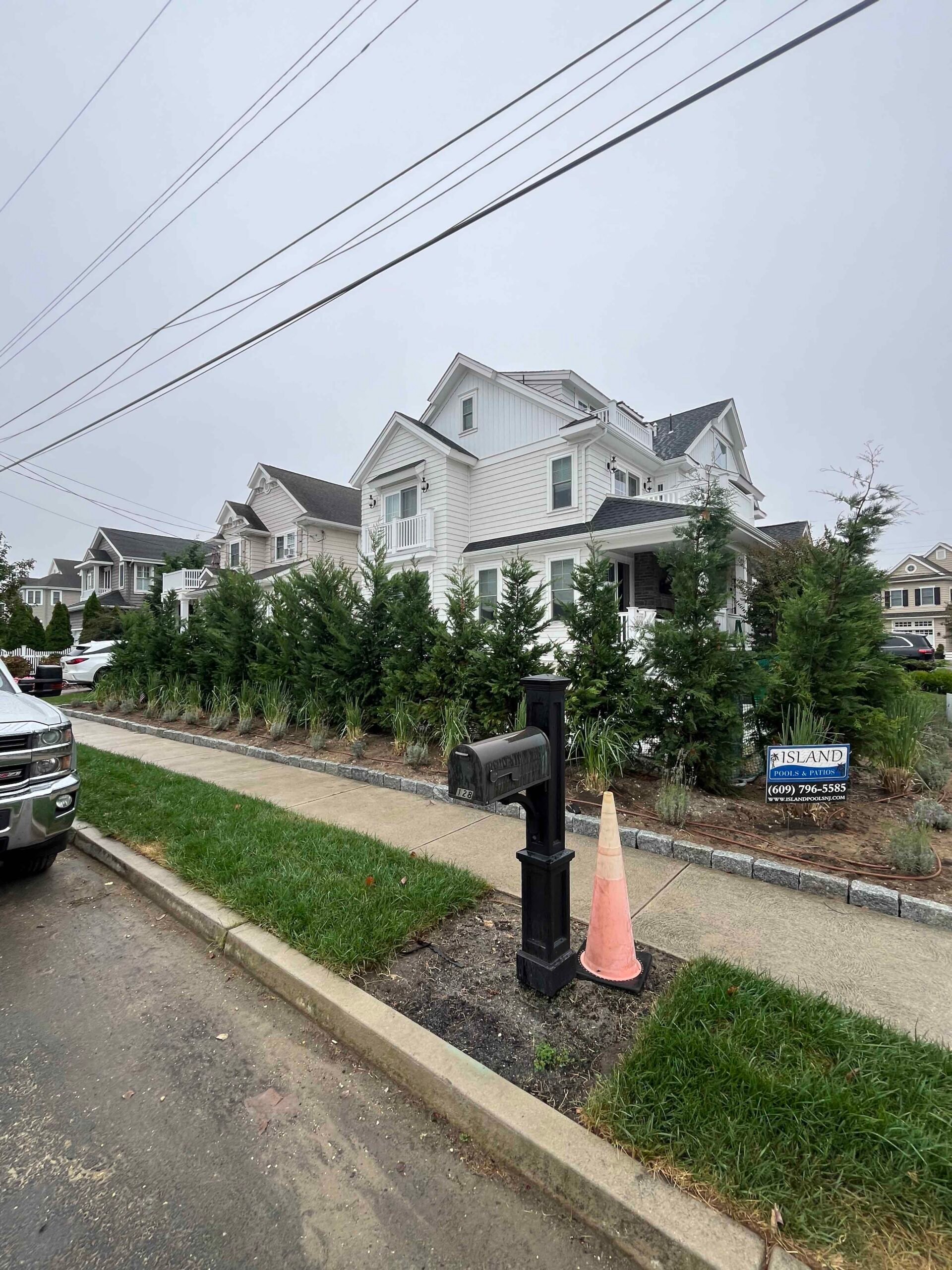 A dense row of newly planted arborvitae lines the front property border of a white residential home, creating a privacy screen along the sidewalk. The bed is freshly graded with dark bare soil, and a traffic cone marks a newly installed black mailbox post 