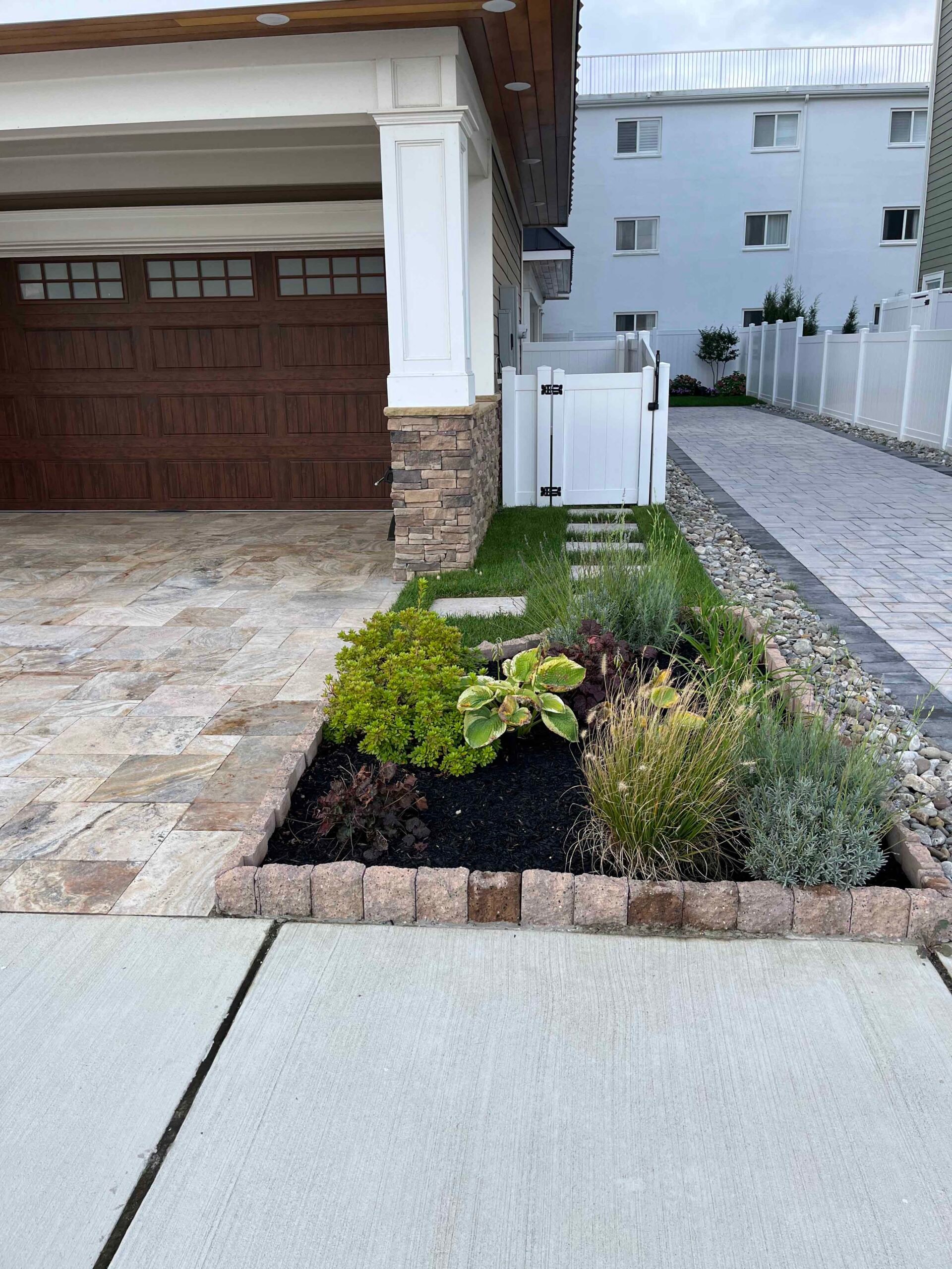 A raised front foundation planting bed with tumbled concrete block edging contains black mulch, hosta, ornamental grasses, sedum, and lavender beside a travertine or natural stone paver driveway; a small turf panel with stepping stones leads to a white vin