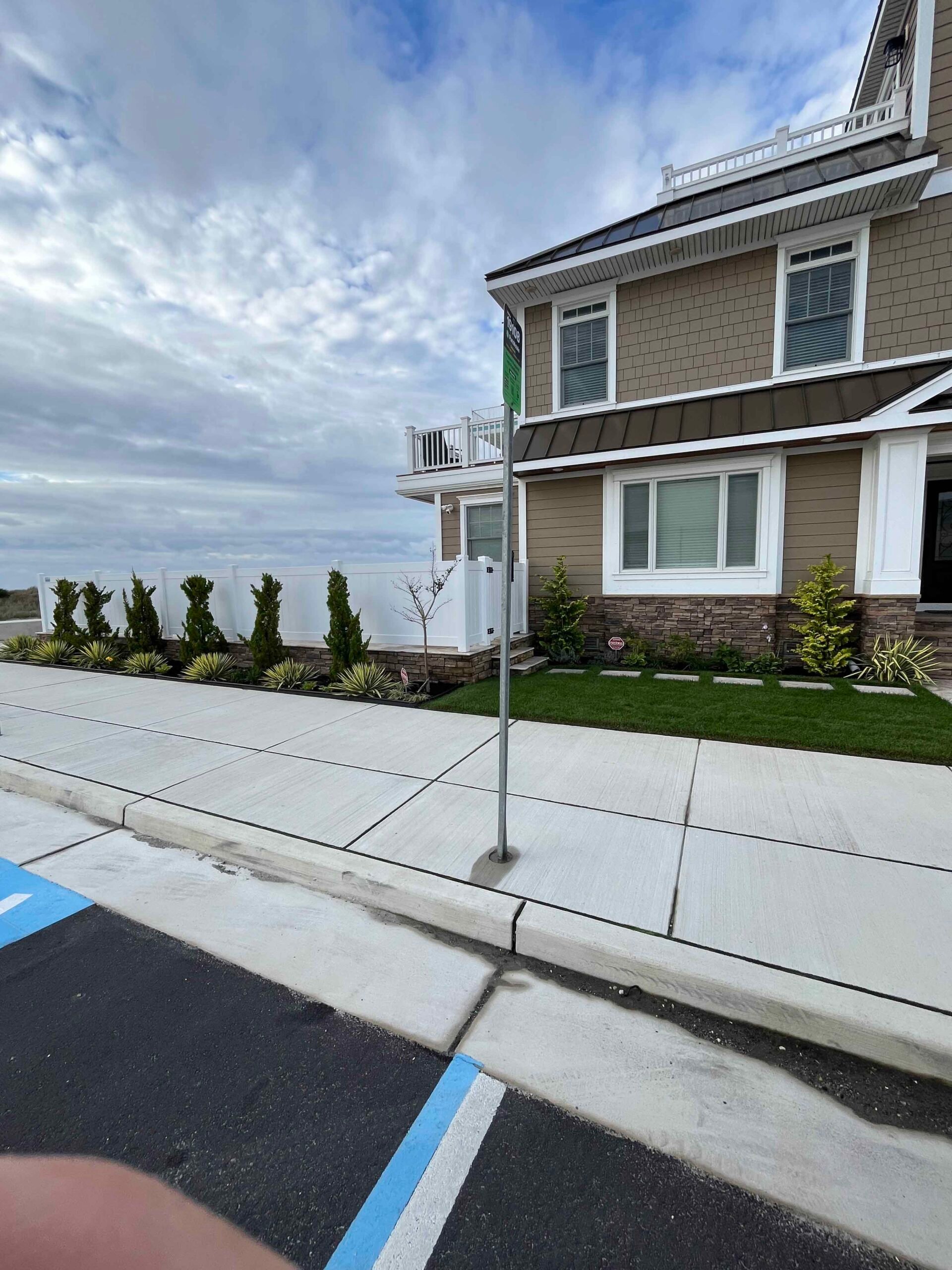 Newly installed foundation planting beds along a commercial building feature columnar arborvitae, ornamental grasses, and small deciduous trees set in a strip of turf beside a concrete sidewalk and parking lot.