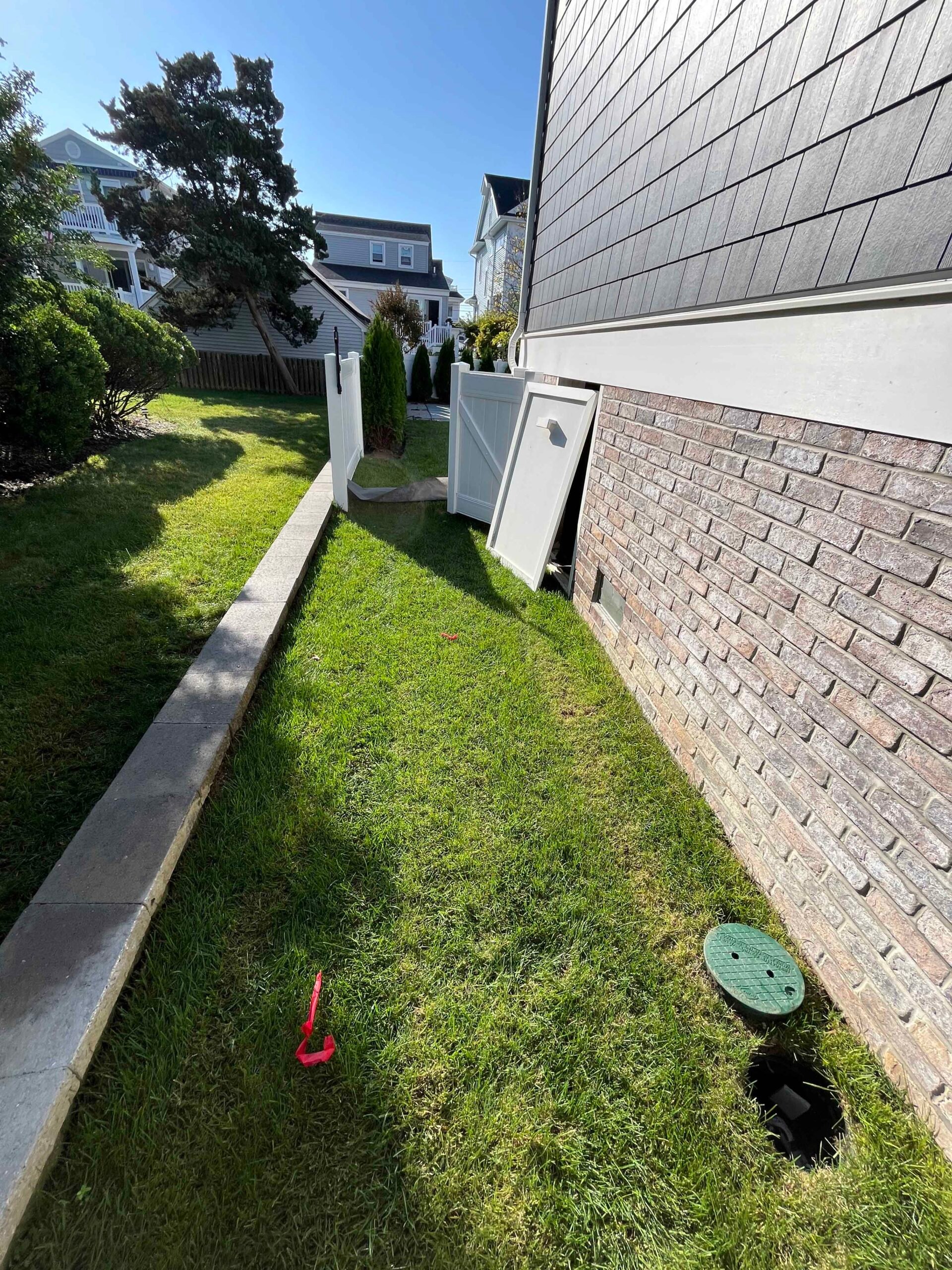 A narrow side yard strip of freshly maintained green turf runs between a brick foundation wall and a concrete mow edge, with a white vinyl privacy fence and columnar evergreen shrubs visible at the far end.