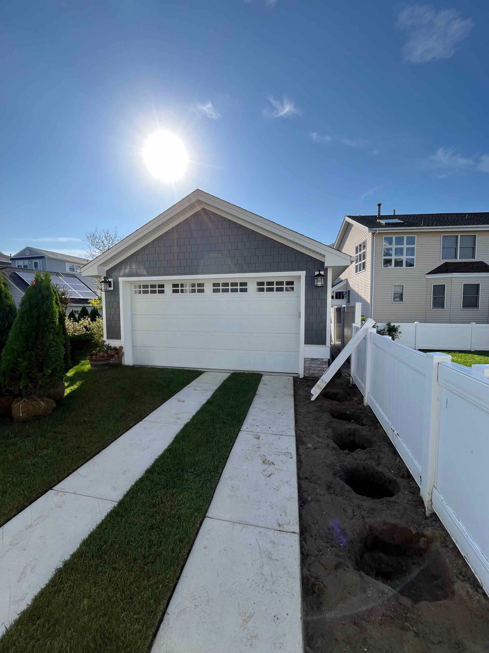 A newly constructed driveway consists of two parallel concrete runners with a strip of freshly sodded turf between them leading to a gray garage. Along the right side, a planting bed is prepared with evenly spaced holes dug in bare soil, ready for plant in