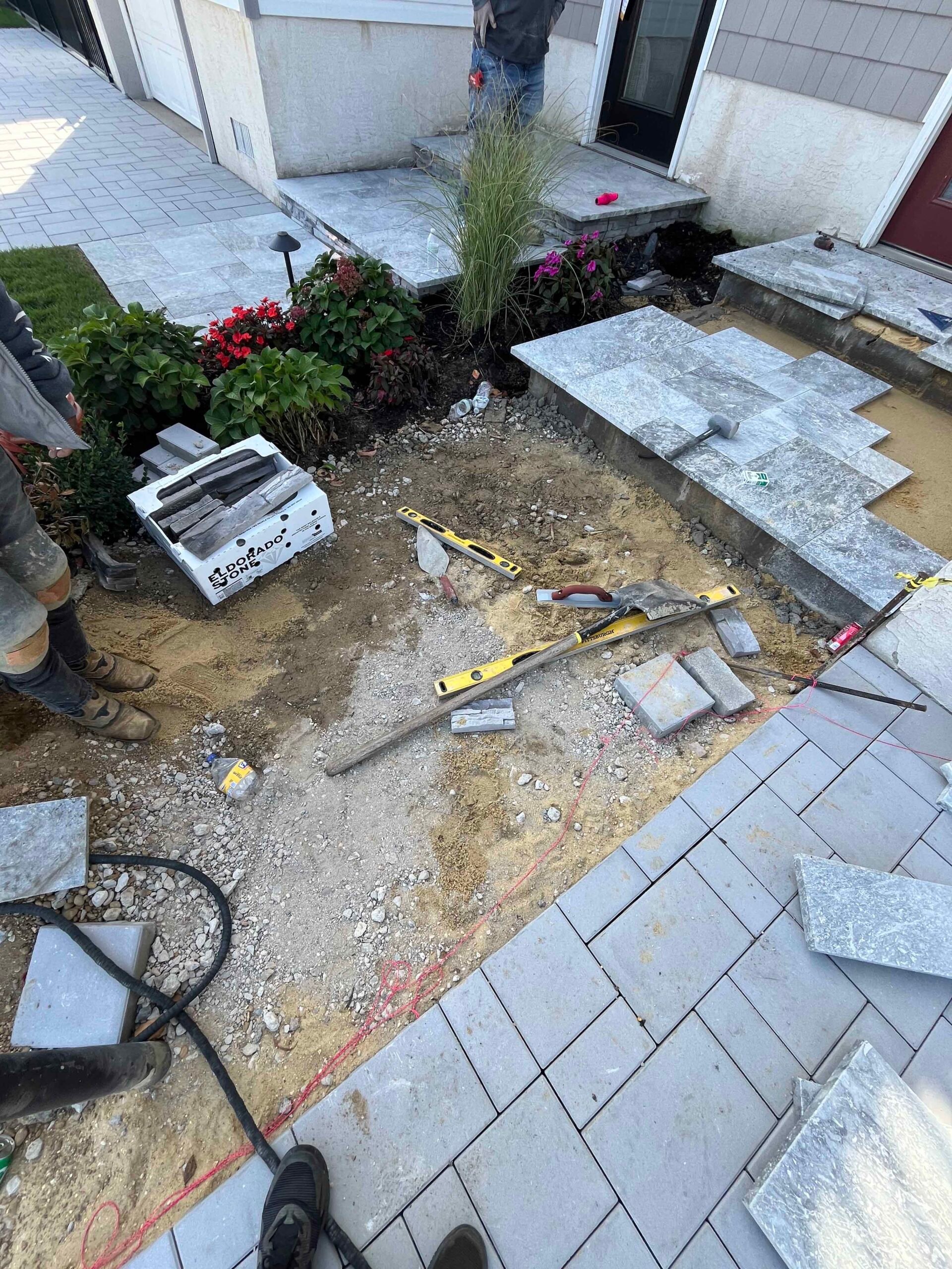 A driveway or walkway area mid-installation showing granite pavers partially laid alongside exposed gravel base, with a spirit level and hand tools on the ground. Existing planting bed with ornamental grasses, red impatiens, and pink flowers is visible at 
