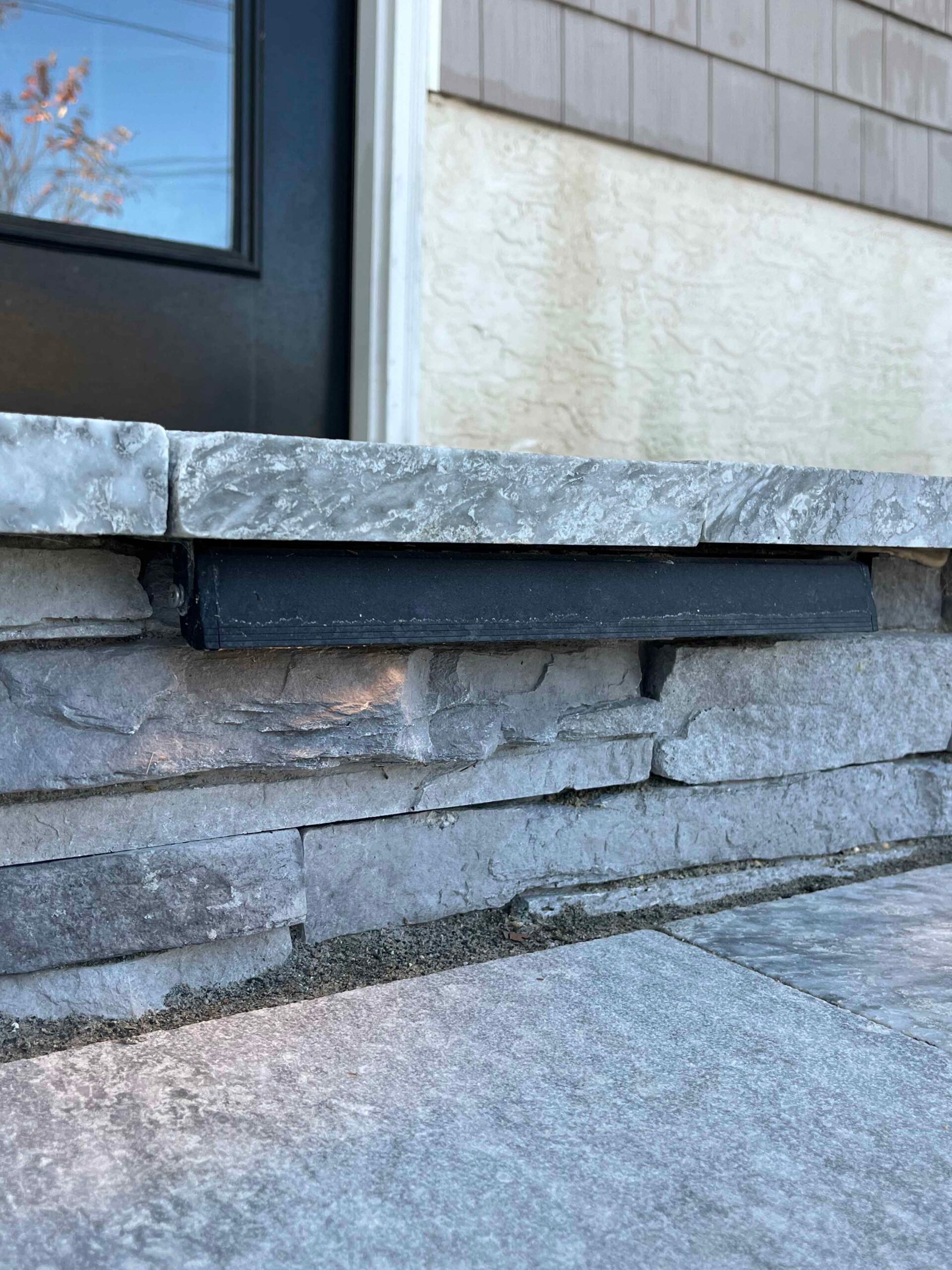 A close-up of a dry-stacked natural bluestone-gray ledgestone retaining or entry wall with a black rectangular recessed step light mounted under the stone cap, adjacent to a bluestone paver surface.