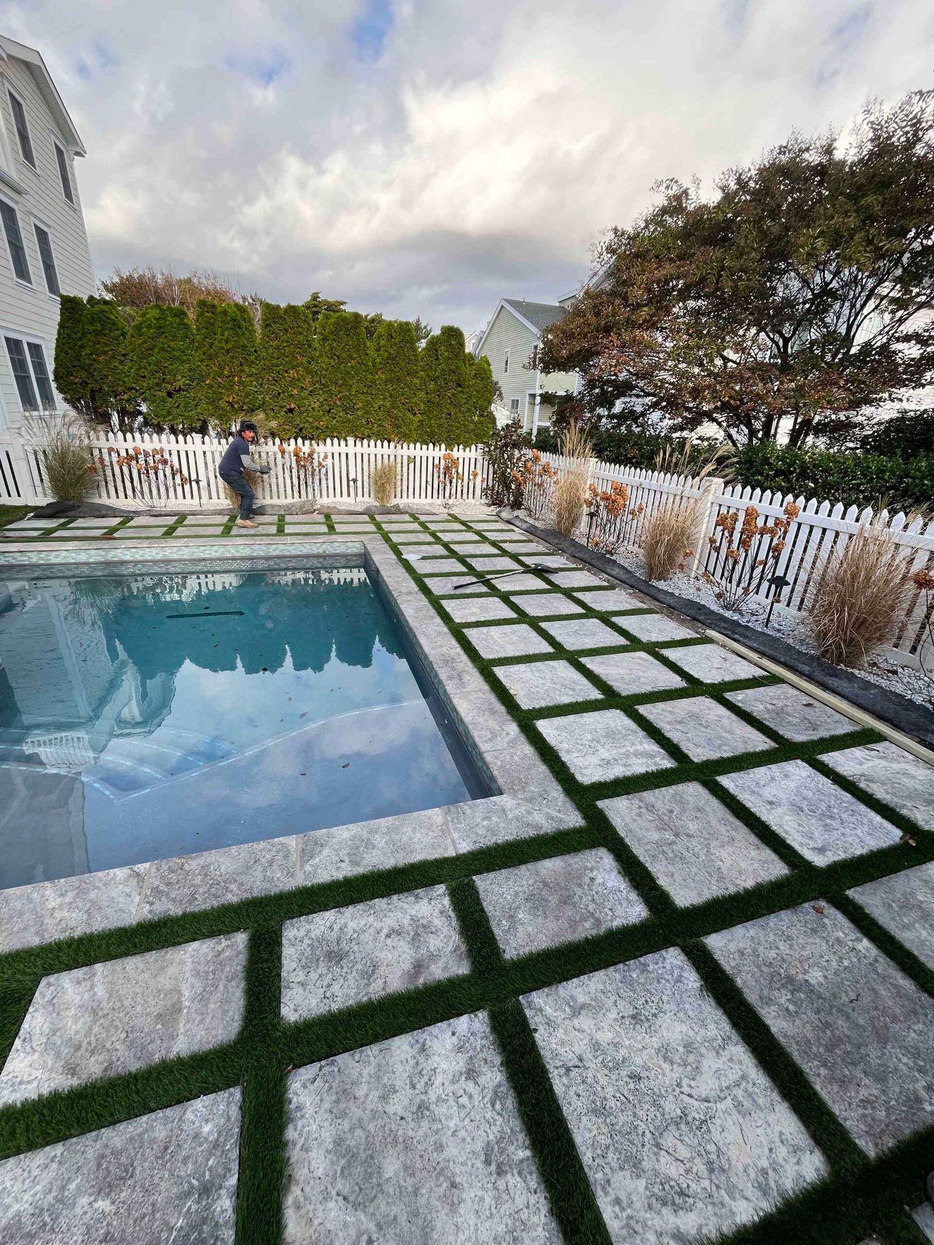 A rectangular in-ground pool is surrounded by large silver-gray natural stone pavers set with artificial turf joints, backed by a tall arborvitae hedge and ornamental grasses along a white picket fence.