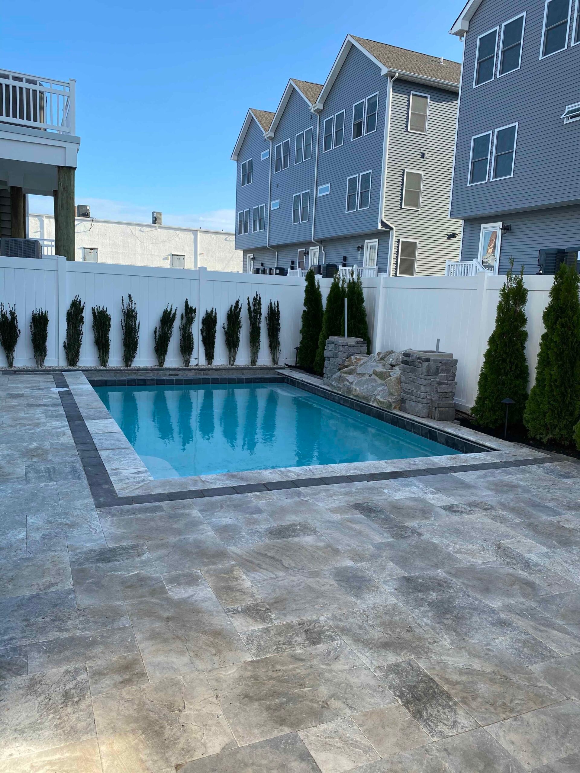 A rectangular in-ground pool is surrounded by large-format tumbled travertine pavers in a silver-gray tone. A row of columnar arborvitae lines the white vinyl privacy fence at the rear, and a natural stone accent wall anchors one corner of the pool surroun