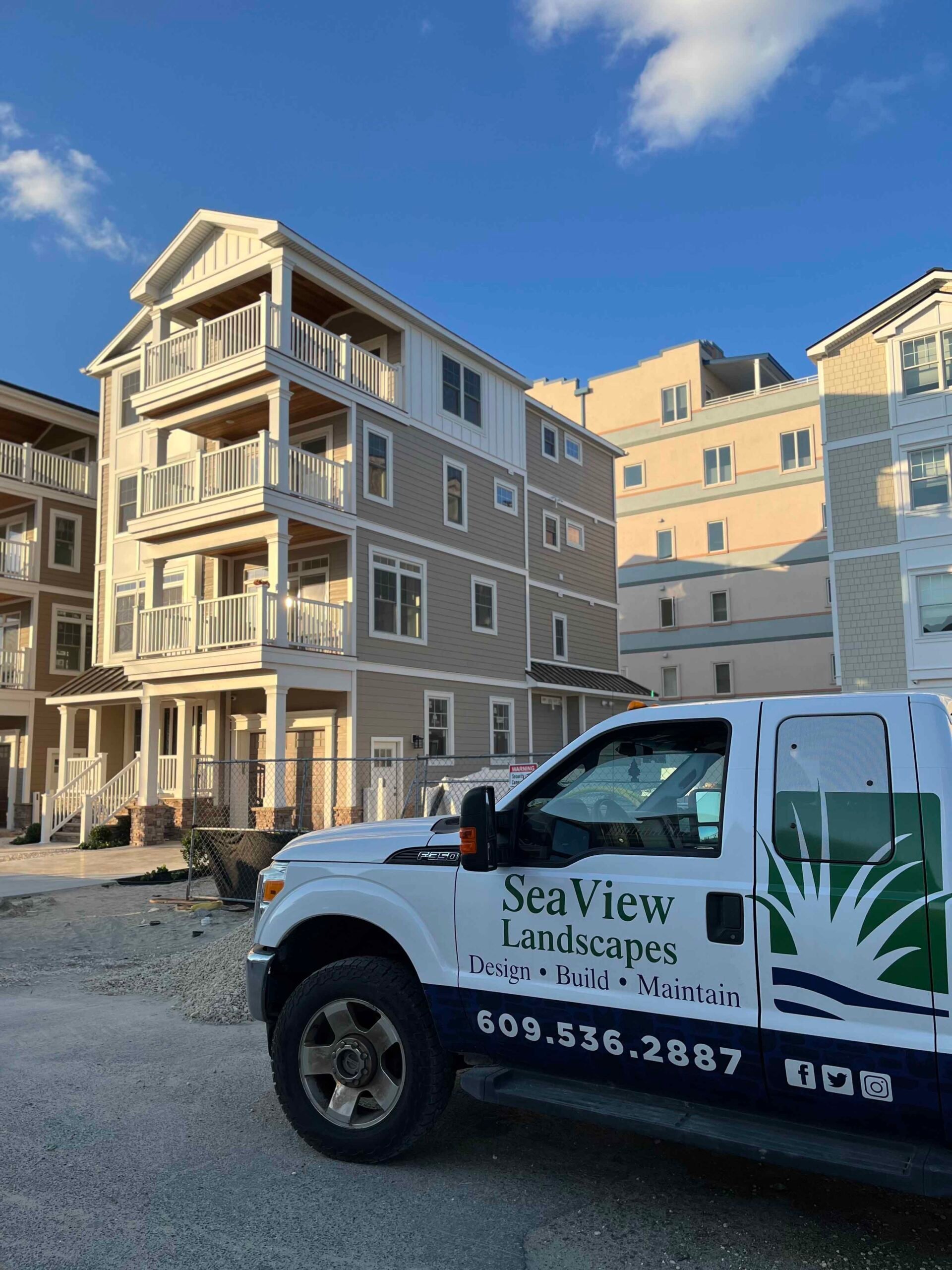 A SeaView Landscapes branded pickup truck is parked on a gravel surface in front of a newly built multi-story residential building. No plantings are present, indicating a pre-installation site visit at an active construction project.