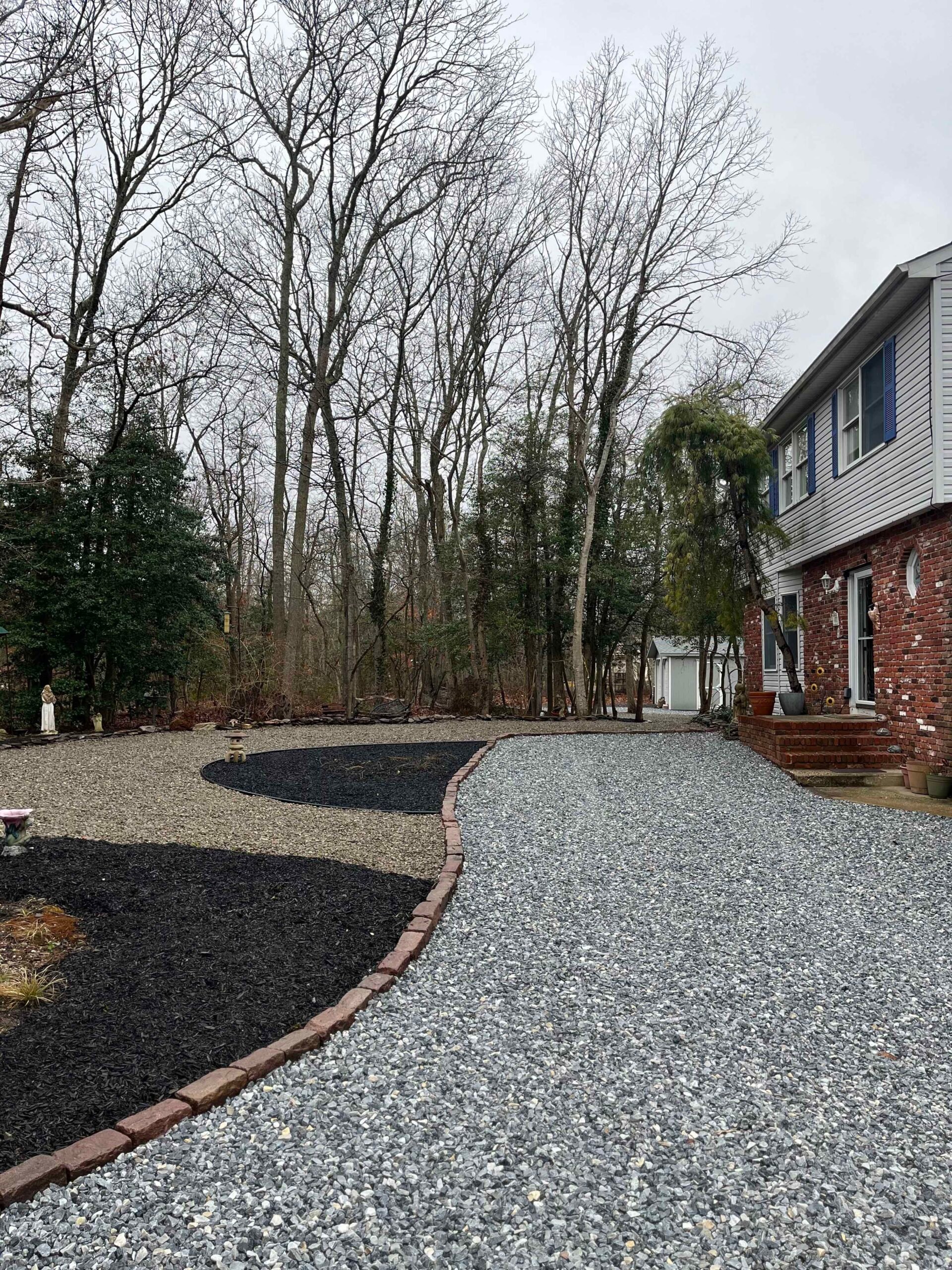 A wide gravel driveway runs along a residential property with a curved black mulch planting bed edged in brick pavers along the left side. Bare deciduous trees fill the background, indicating a late fall or winter season.