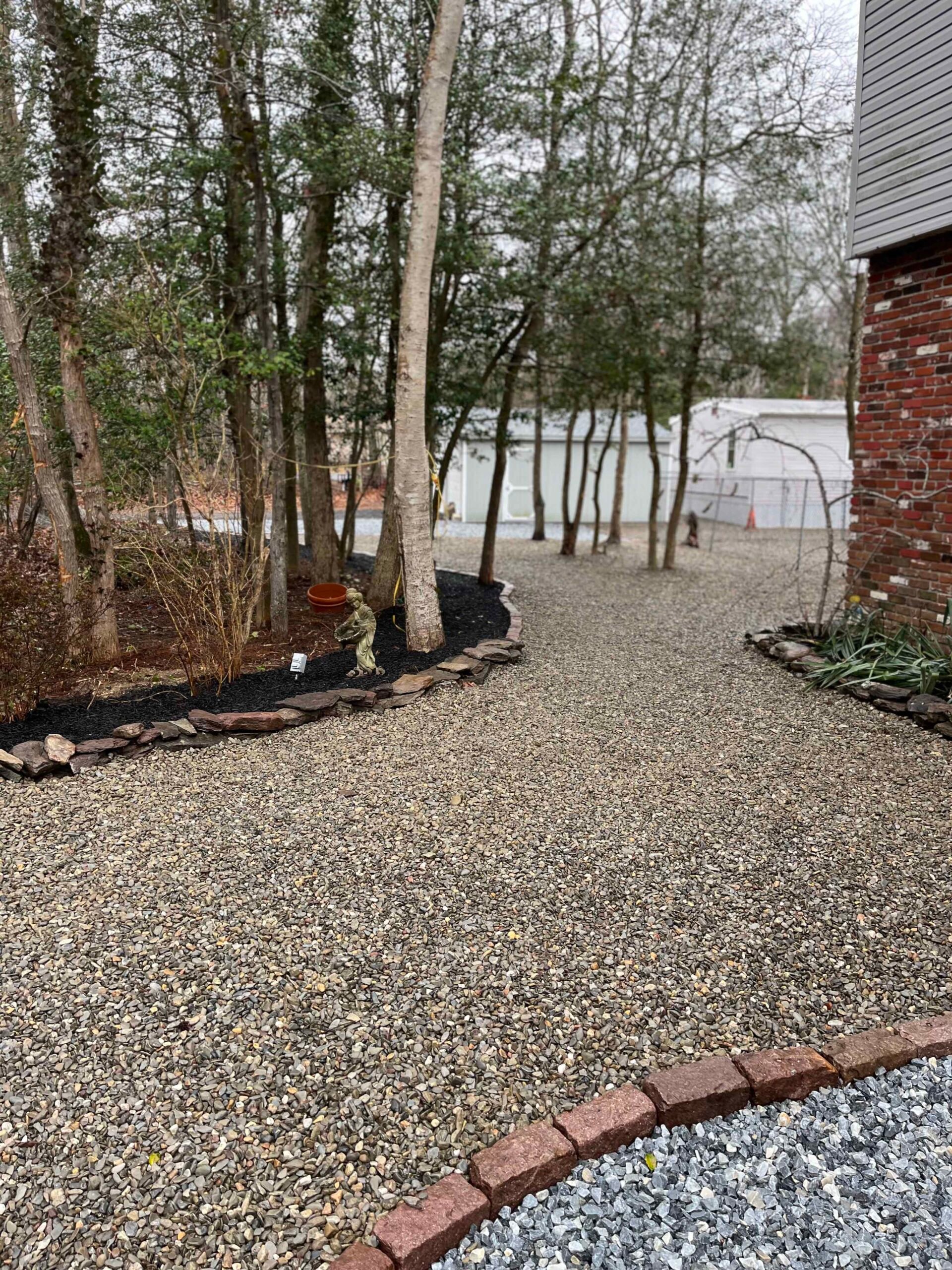 A wide gravel pathway made of pea gravel or crushed stone runs through a wooded side yard, flanked by paver brick edging and dark mulch tree ring beds around birch or multi-trunk deciduous trees. Small planting beds with early spring bulb growth are visibl