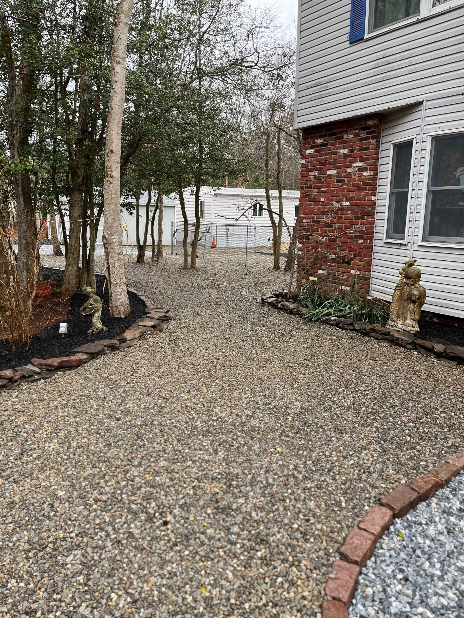 A crushed gravel walkway runs between the side of a brick-accented house and a row of deciduous and evergreen trees, bordered on both sides by flat brick edging. The tree bases have dark mulch rings. Two stone garden statues flank the path. Bare tree canop
